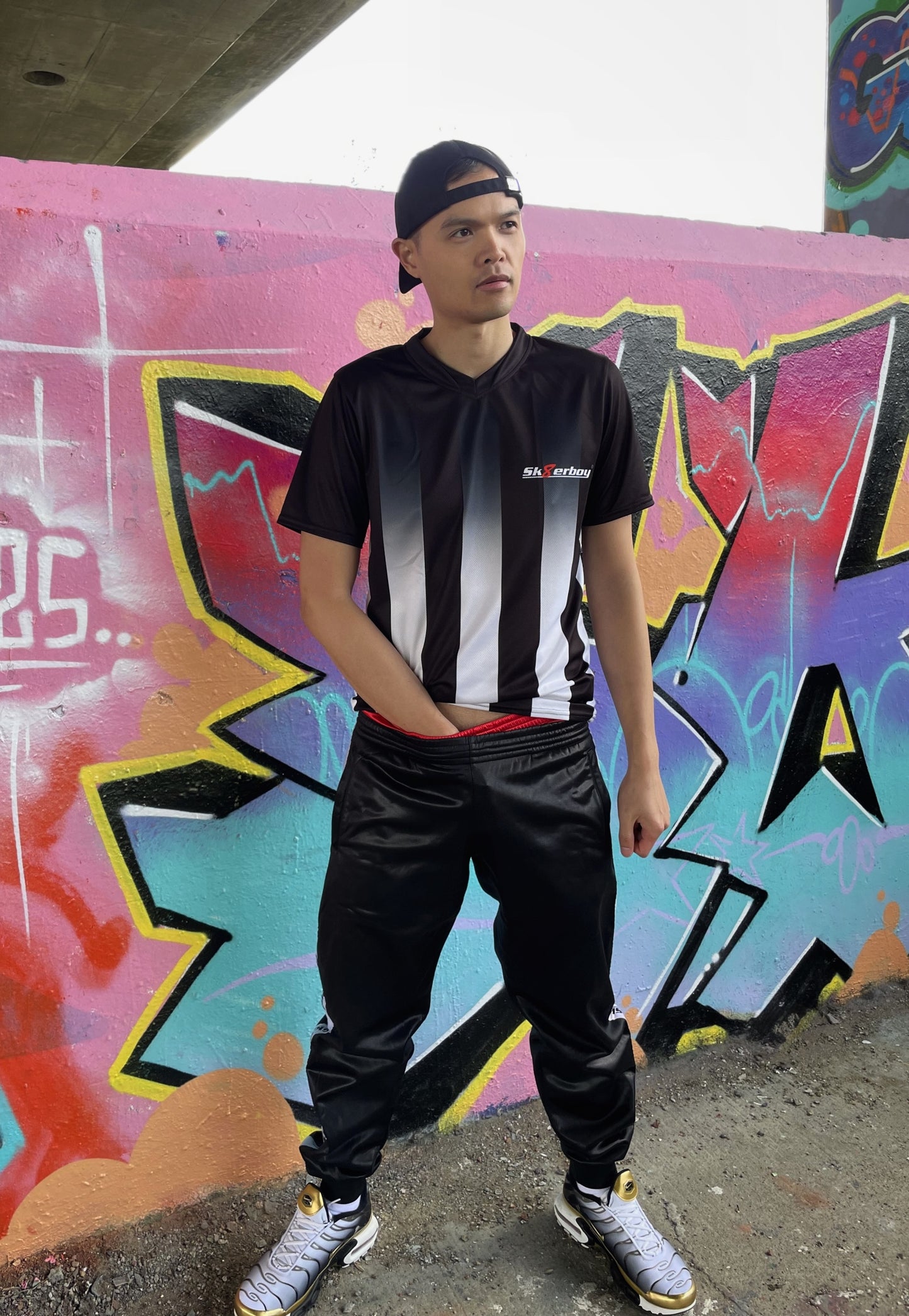 Model trägt das Sk8erboy® Tikot in Schwarz-Weiß mit vertikalen Streifen vor bunter Graffiti-Wand und zeigt auf das Logo auf der Brust.
