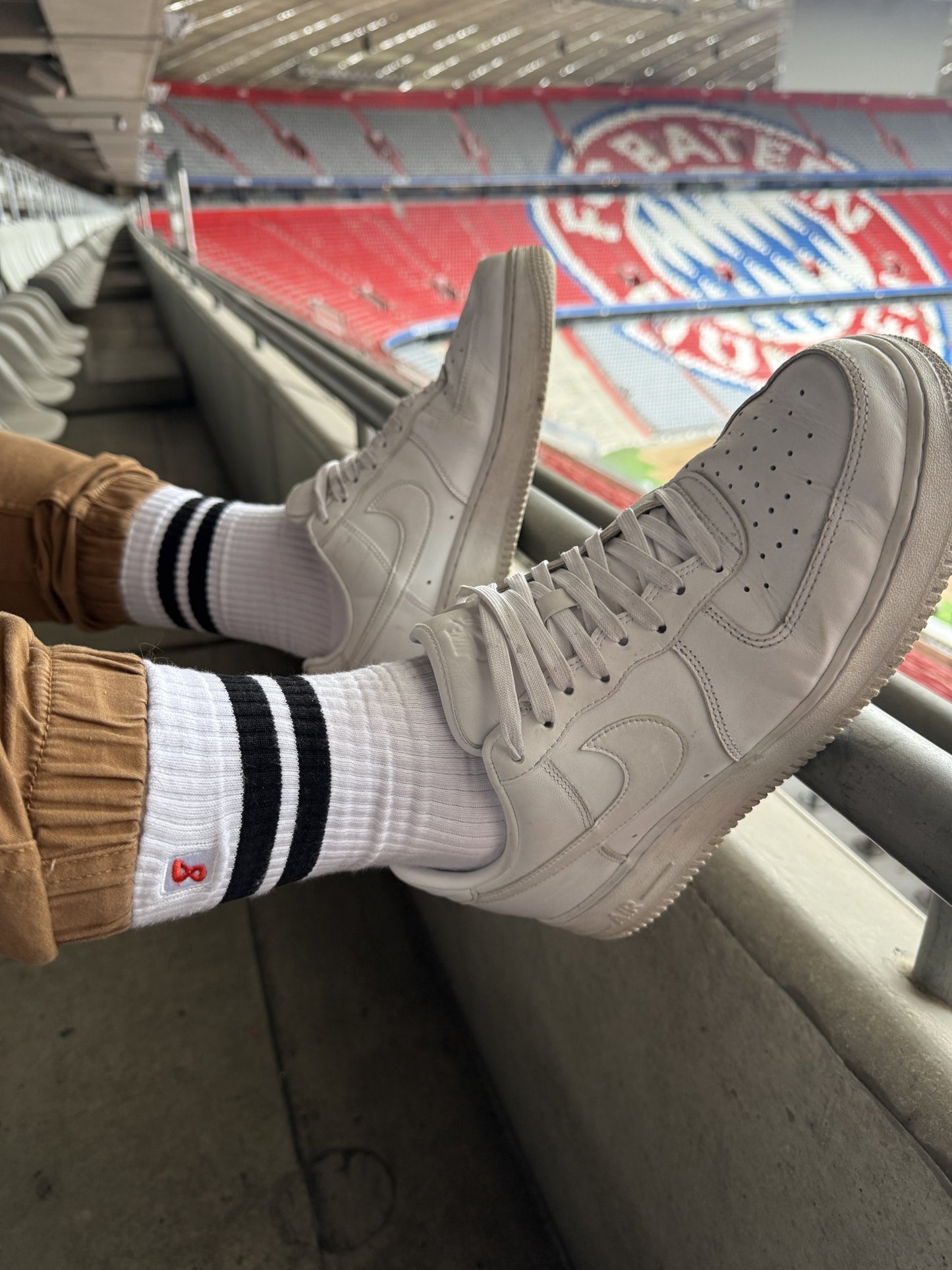 Nahaufnahme von Sk8erboy® Retro Socken mit schwarzen Streifen, getragen mit weißen Nike-Sneakern im Stadion Allianz Arena München FC Bayern sportlicher Gay-Look mit Statement-Faktor.