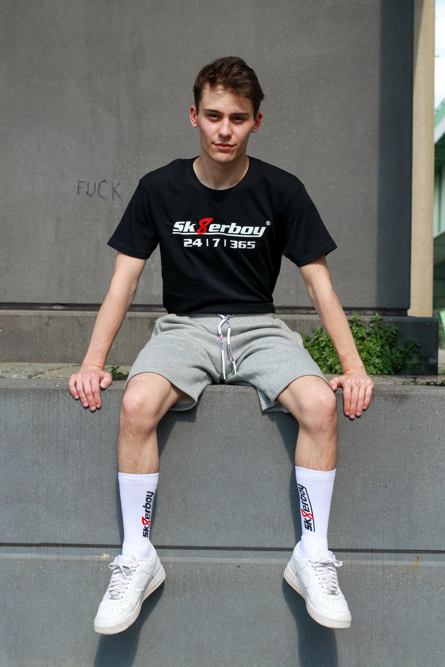 hübscher gay twink trägt ein sk8erboy t-shirt shirt in schwarz baumwolle cotton mit einem sk8erboy aufdruck mit weisser schrift und 8 acht in rot mit 24 7 365 fetter aufdruck mit dem trainingsanzug in kurz und grau von sk8erboy