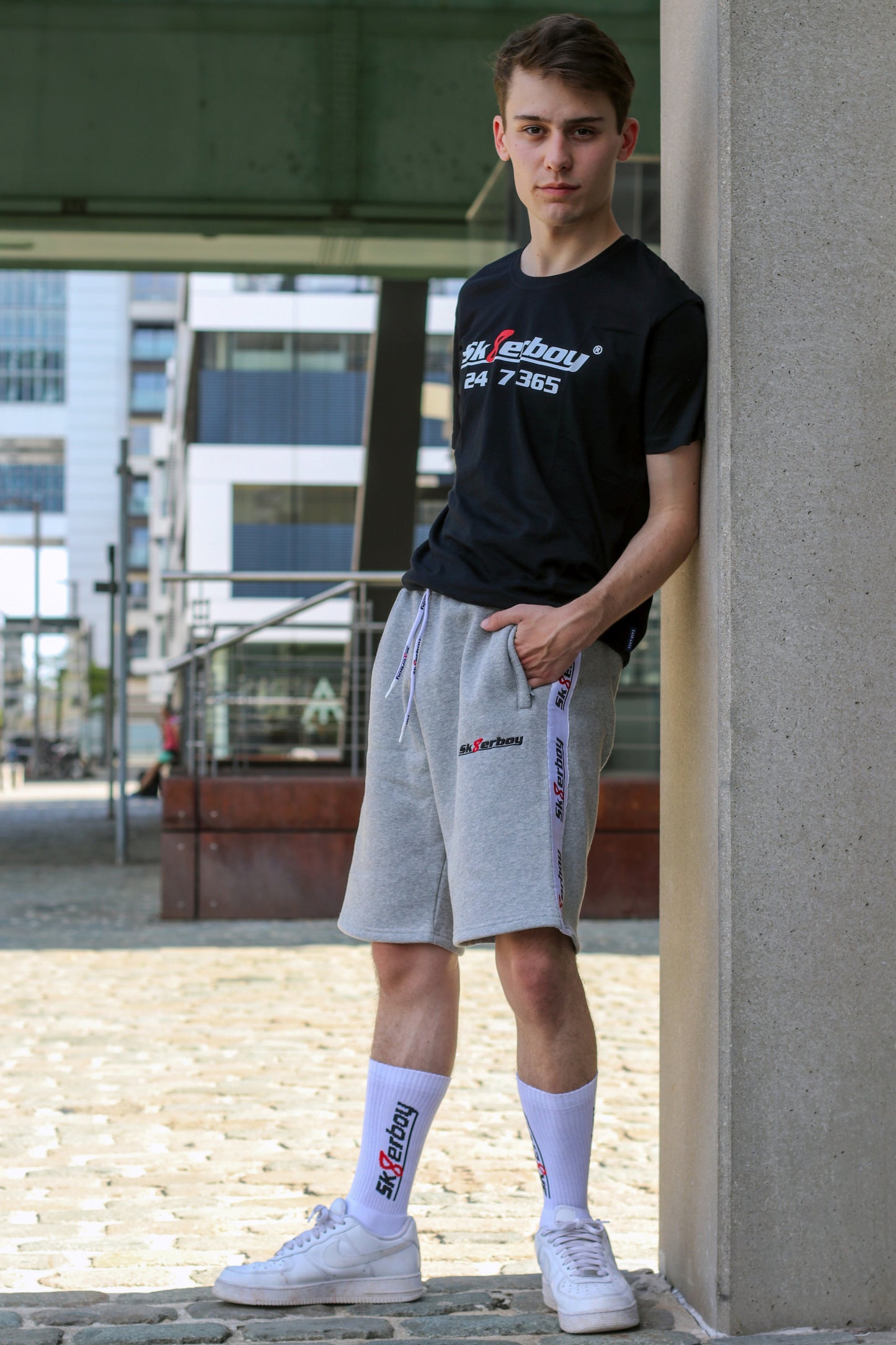hübscher gay twink trägt ein sk8erboy t-shirt shirt in schwarz baumwolle cotton mit einem sk8erboy aufdruck mit weisser schrift und 8 acht in rot mit 24 7 365 fetter aufdruck mit dem trainingsanzug in kurz und grau von sk8erboy