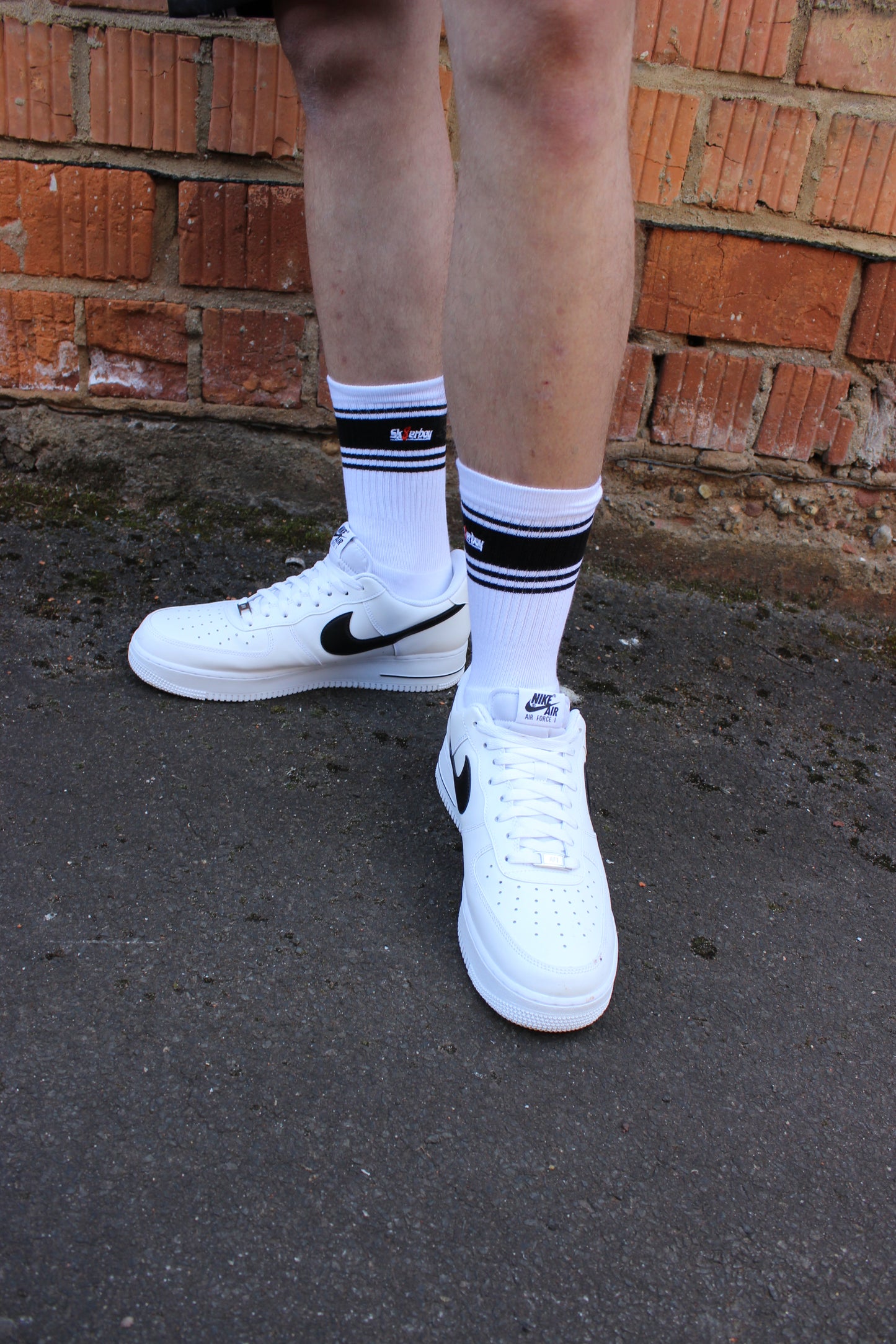 sk8erboy deluxe socken sox in weiss mit schwarzen ringen schwarzen mit schwarzem streifen und logo einstickung rote 8 acht er traegt nike afo air force one low mit weissem leder und schwarzem leder nike swoosh haken  im hintergrund ein lostplace der boy hat einen blanken unterschenkel mit haaren