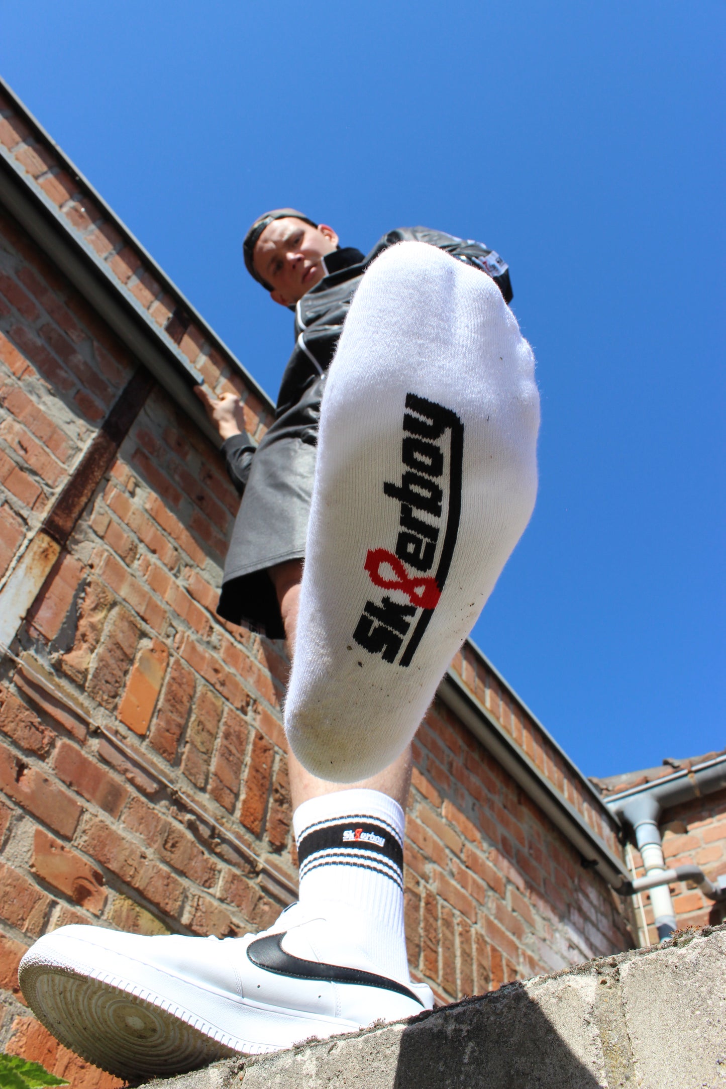 sk8erboy deluxe socken sox in weiss mit schwarzen ringen schwarzen mit schwarzem streifen und logo einstickung rote 8 acht er traegt nike afo air force one low mit weissem leder und schwarzem leder nike swoosh haken im hintergrund ein lostplace der boy hat einen blanken unterschenkel mit haaren