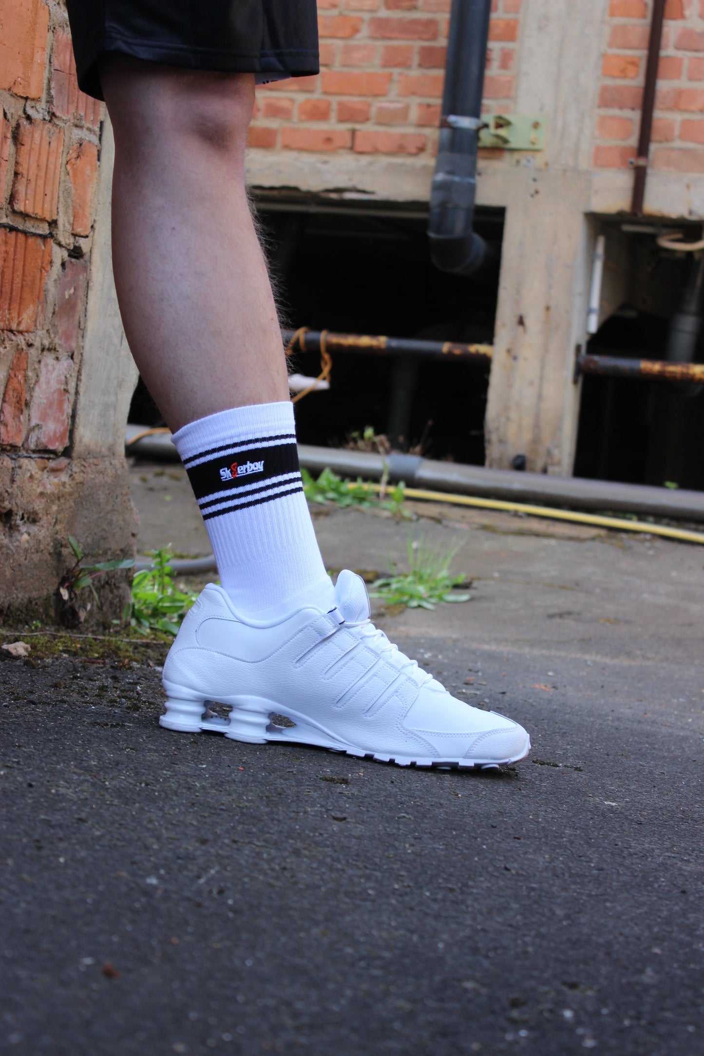sk8erboy deluxe socken sox in weiss mit schwarzen ringen schwarzen mit schwarzem streifen und logo einstickung rote 8 acht er traegt nike afo air force one low mit weissem leder und schwarzem leder nike swoosh haken im hintergrund ein lostplace der boy hat einen blanken unterschenkel mit haaren