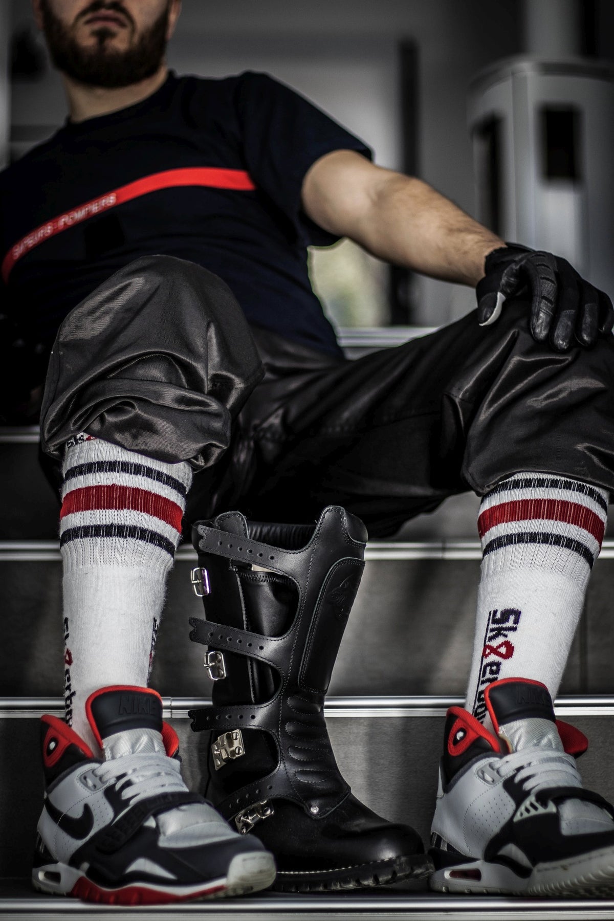 homme gay sportif avec des gants en cuir, assis en survêtement sur les marches et portant dans ses sneakers Nike des tubesocks Sk8erboy noirs, blancs et rouges à rayures et avec grand logo