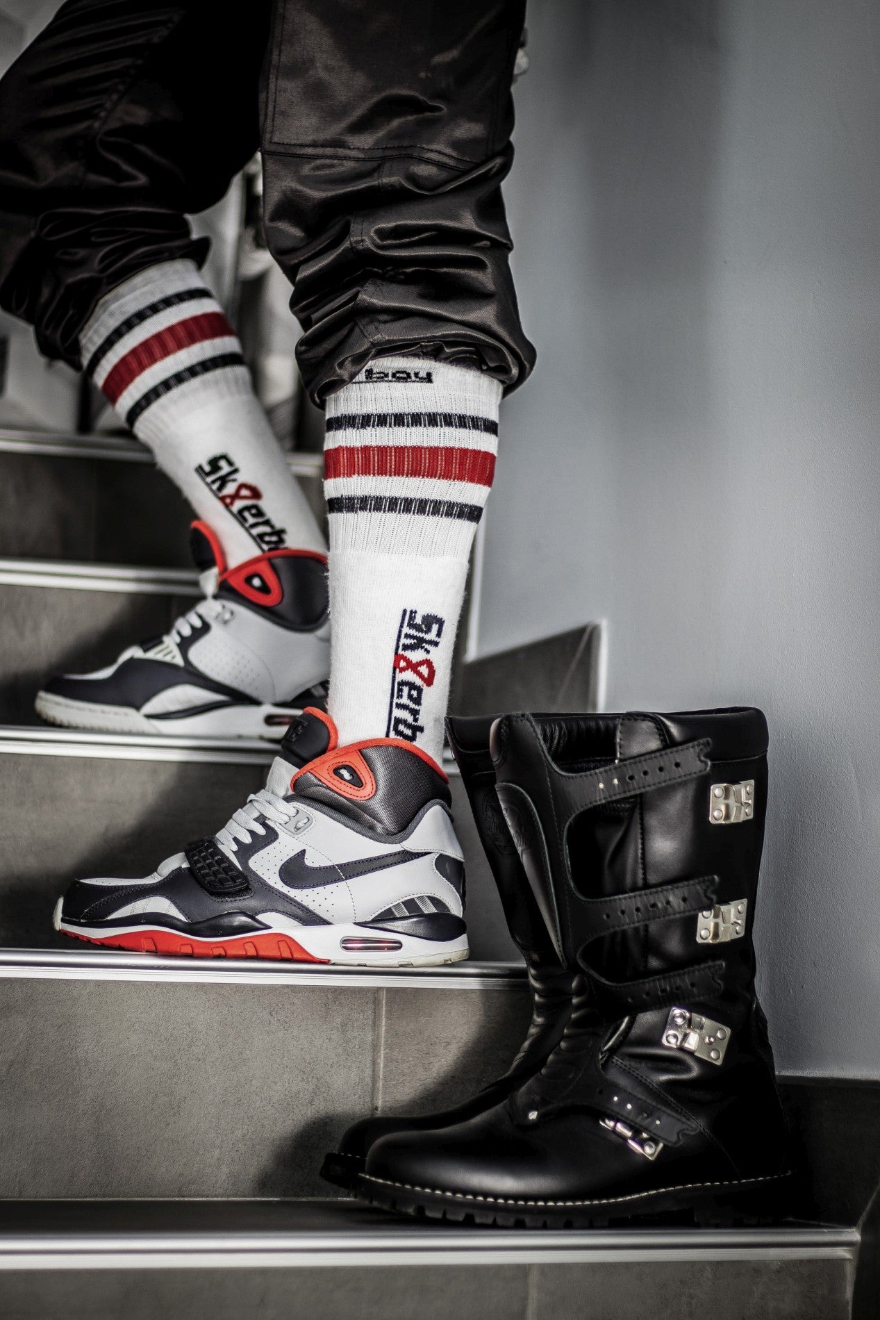 sportif gay portant des sneakers Nike et des chaussettes tubulaires SK8ERBOY avec grand logo sur le côté, pantalon retroussé. Au premier plan, bottes de moto en cuir