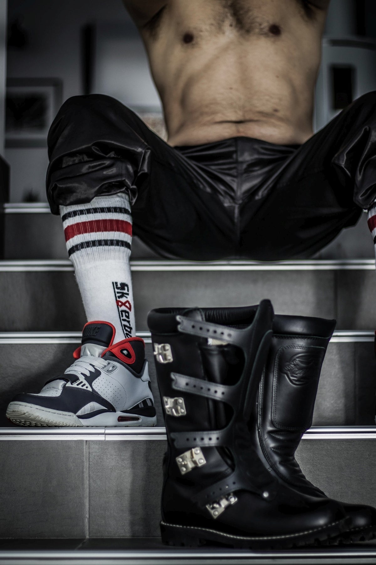 Un homme gay sportif est assis sur un escalier et porte dans ses sneakers des chaussettes Sk8erboy tube noires, blanches et rouges avec grand logo sur le côté, et des bottes biker en cuir devant lui.