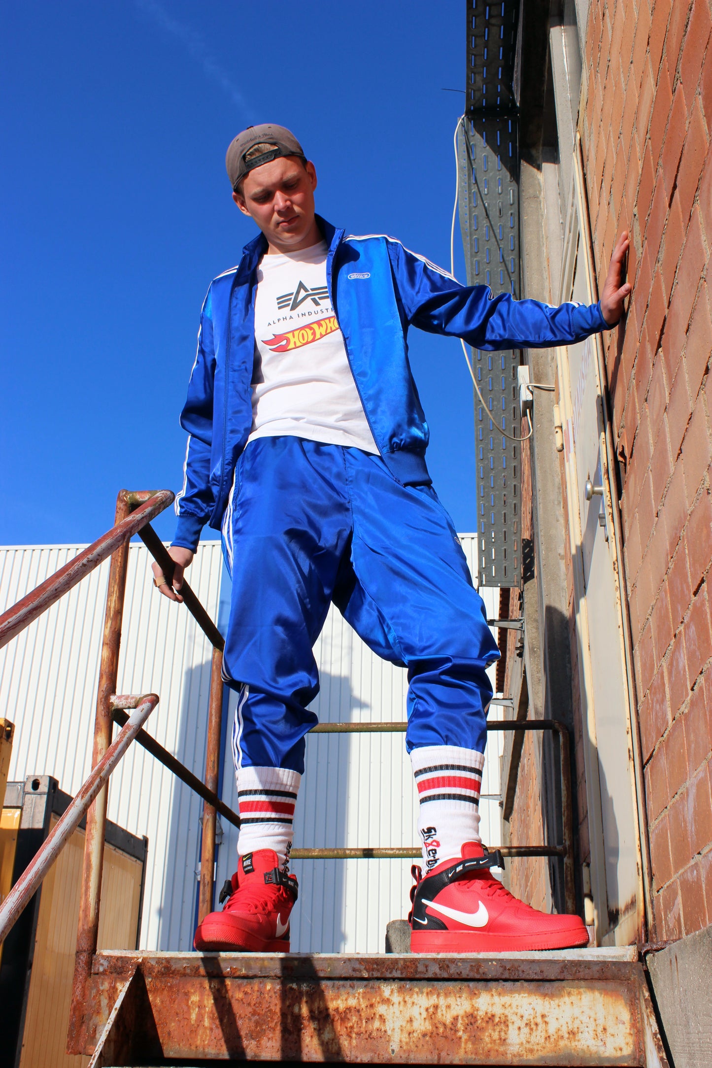 sk8erboy chaussettes tube sans talon dans une Nike Air Force Mid rouge en cuir avec boucle noire et swoosh blanc ; le garçon est sur une grille métallique, porte un pantalon adidas Firebird satiné bleu roi, une veste adidas Firebird brillante bleu roi à bandes blanches, une casquette grise et un T-shirt blanc Alpha Industries avec logo Hot Wheels, se tenant l’entrejambe.