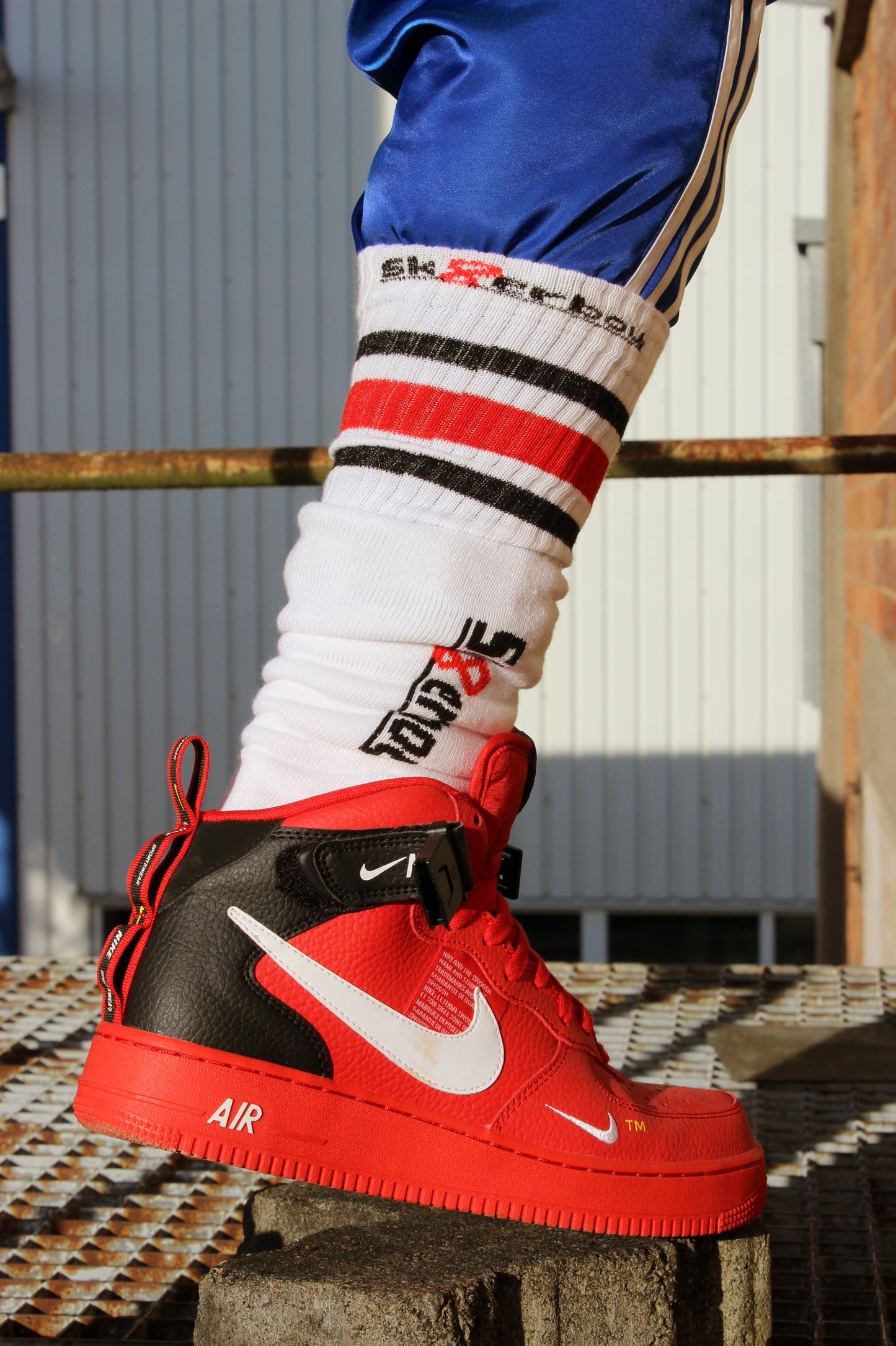 sk8erboy chaussettes tube sans talon, dans une Nike Air Force Mid rouge en cuir avec boucle noire et swoosh blanc. Le garçon est sur une grille métallique, porte un pantalon Adidas Firebird brillant bleu roi, une veste satin Adidas Firebird bleu roi à bandes blanches, une casquette grise et un T-shirt blanc Alpha Industries
