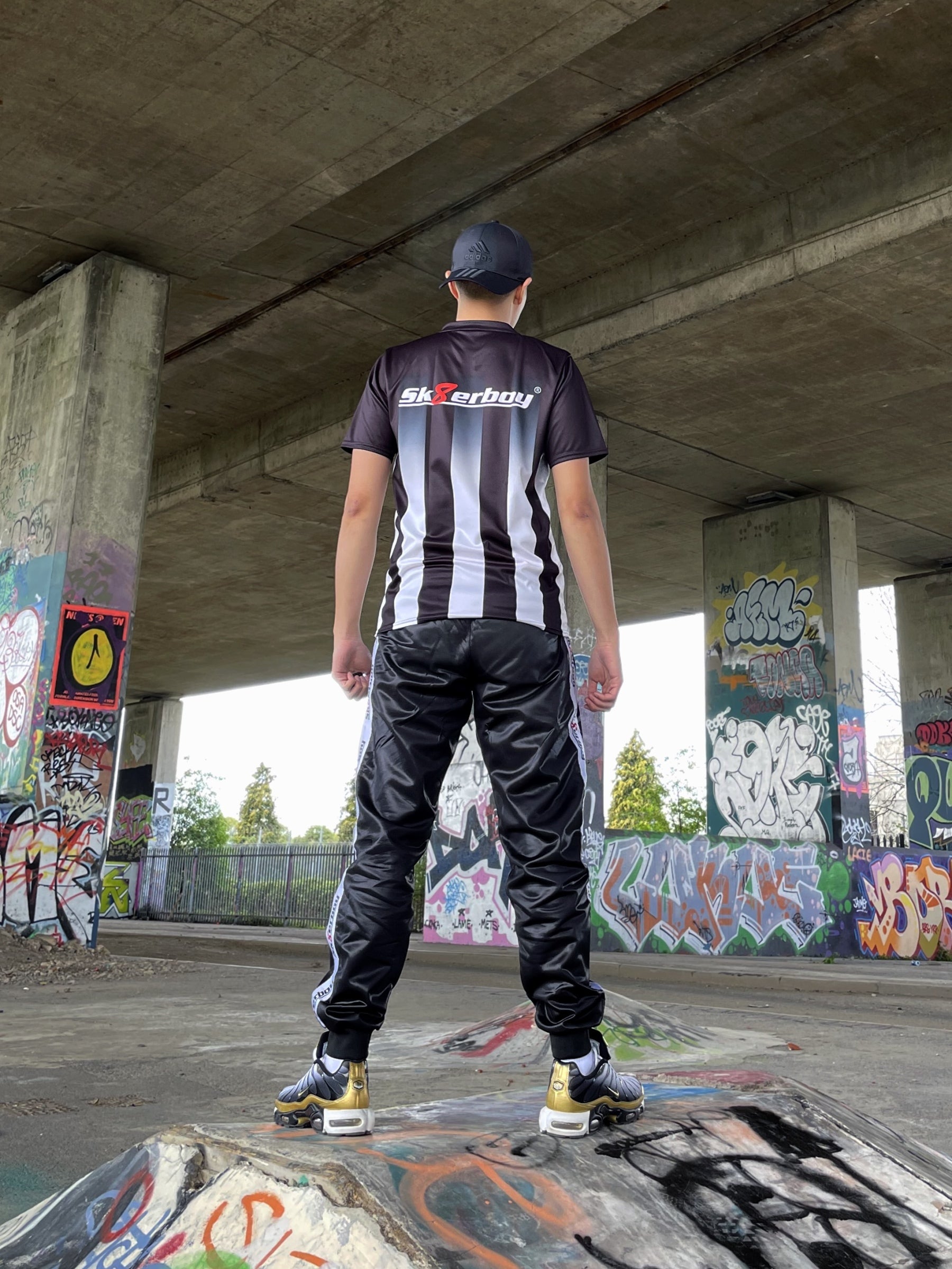 Urbaner Look: Model steht mit dem Sk8erboy® Tikot und glänzender Jogginghose auf einer Halfpipe unter einer Brücke mit Graffiti.
