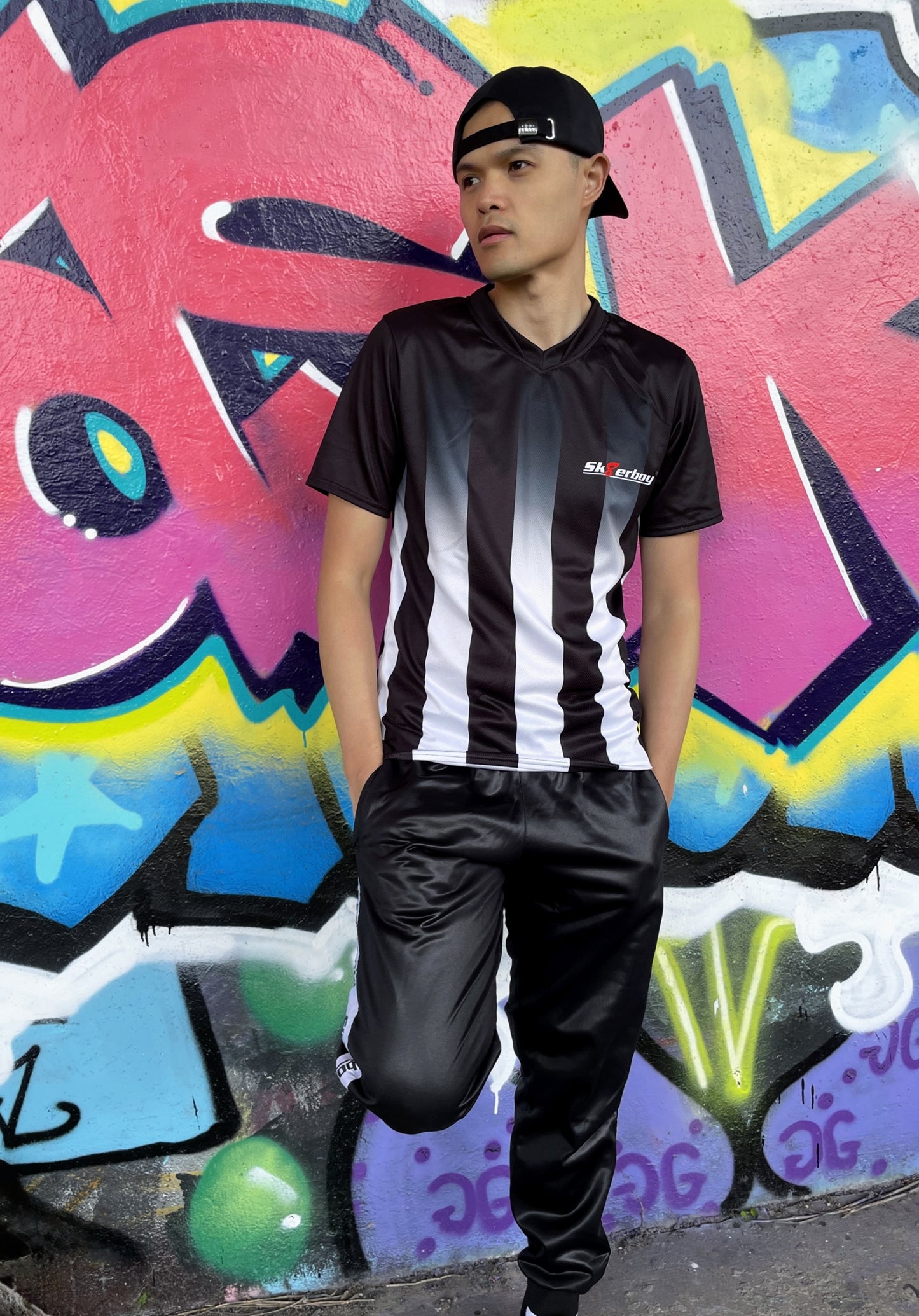 Model trägt das Sk8erboy® Tikot in Schwarz-Weiß mit vertikalen Streifen vor bunter Graffiti-Wand und zeigt auf das Logo auf der Brust.