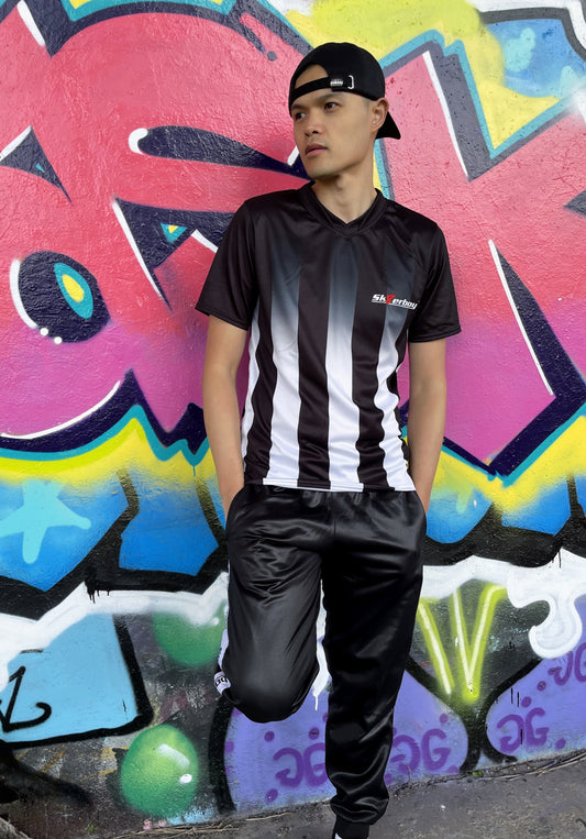 Model trägt das Sk8erboy® Tikot in Schwarz-Weiß mit vertikalen Streifen vor bunter Graffiti-Wand und zeigt auf das Logo auf der Brust.