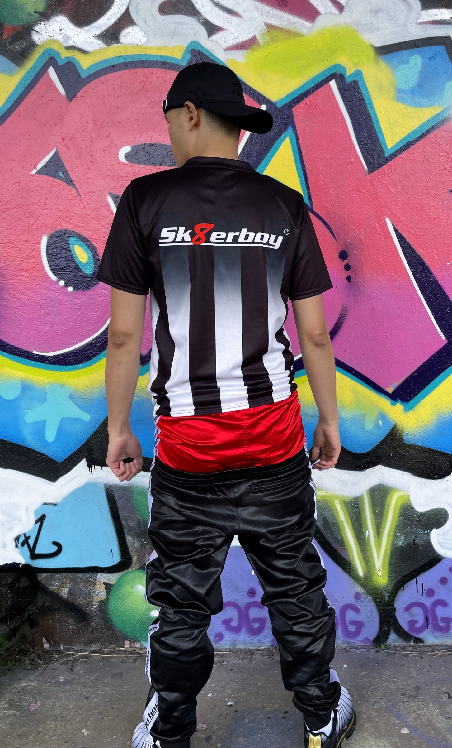 Rückansicht des Sk8erboy® Tikots mit großem Logo, getragen von einem Model in Hocke vor stylischer Graffiti-Kulisse.