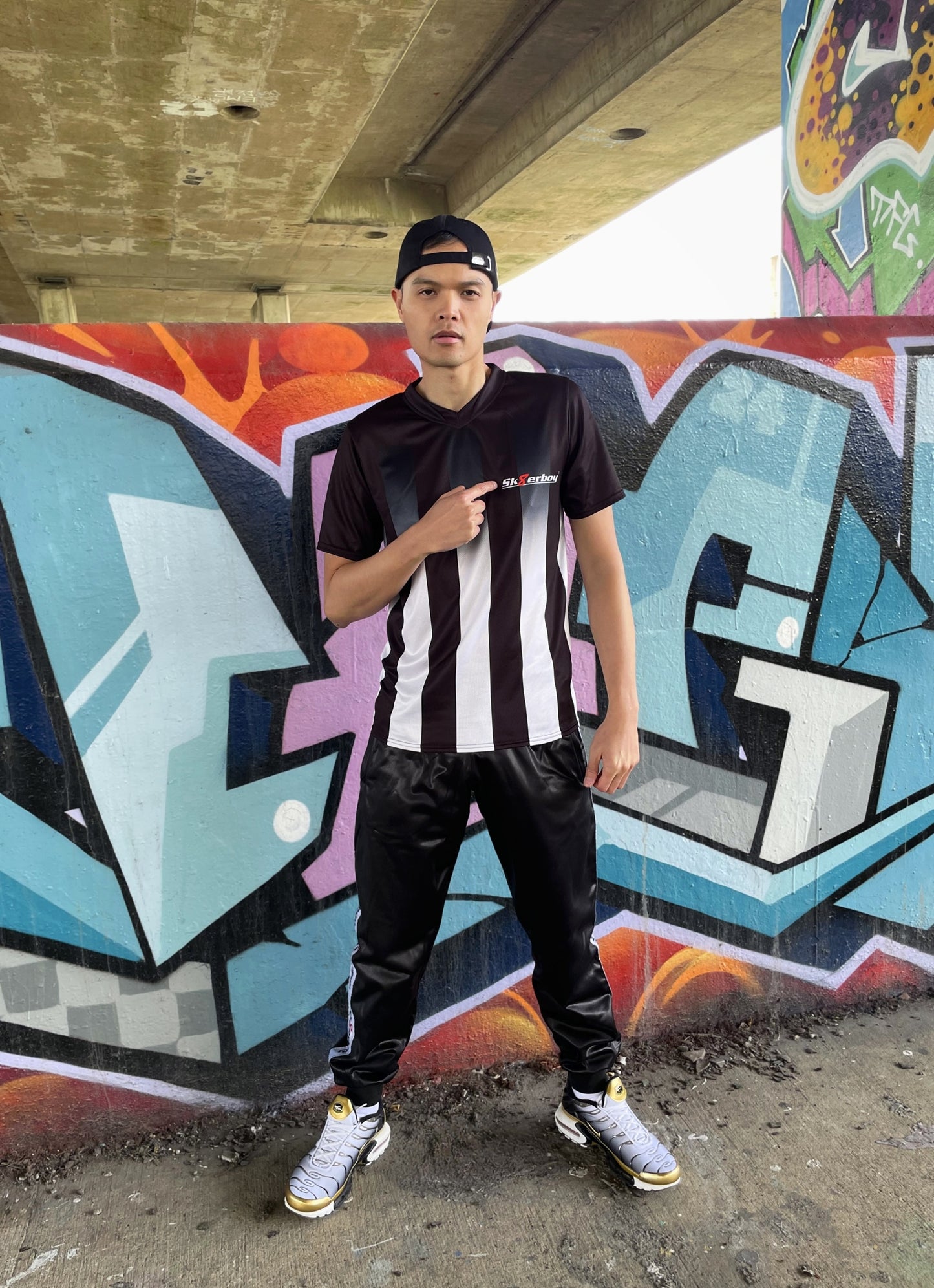 Model trägt das Sk8erboy® Tikot in Schwarz-Weiß mit vertikalen Streifen vor bunter Graffiti-Wand und zeigt auf das Logo auf der Brust.