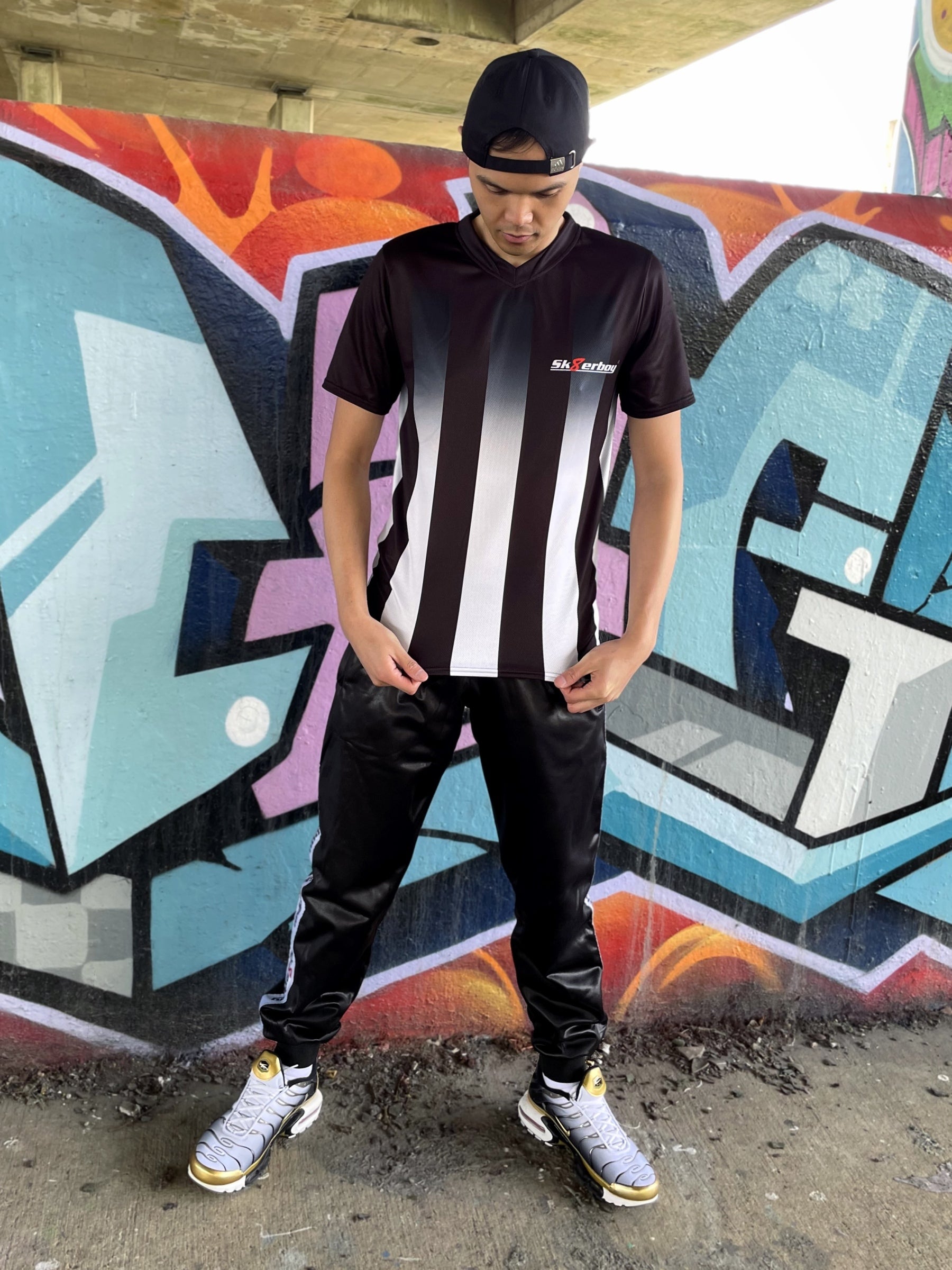 Model trägt das Sk8erboy® Tikot in Schwarz-Weiß mit vertikalen Streifen vor bunter Graffiti-Wand und zeigt auf das Logo auf der Brust.