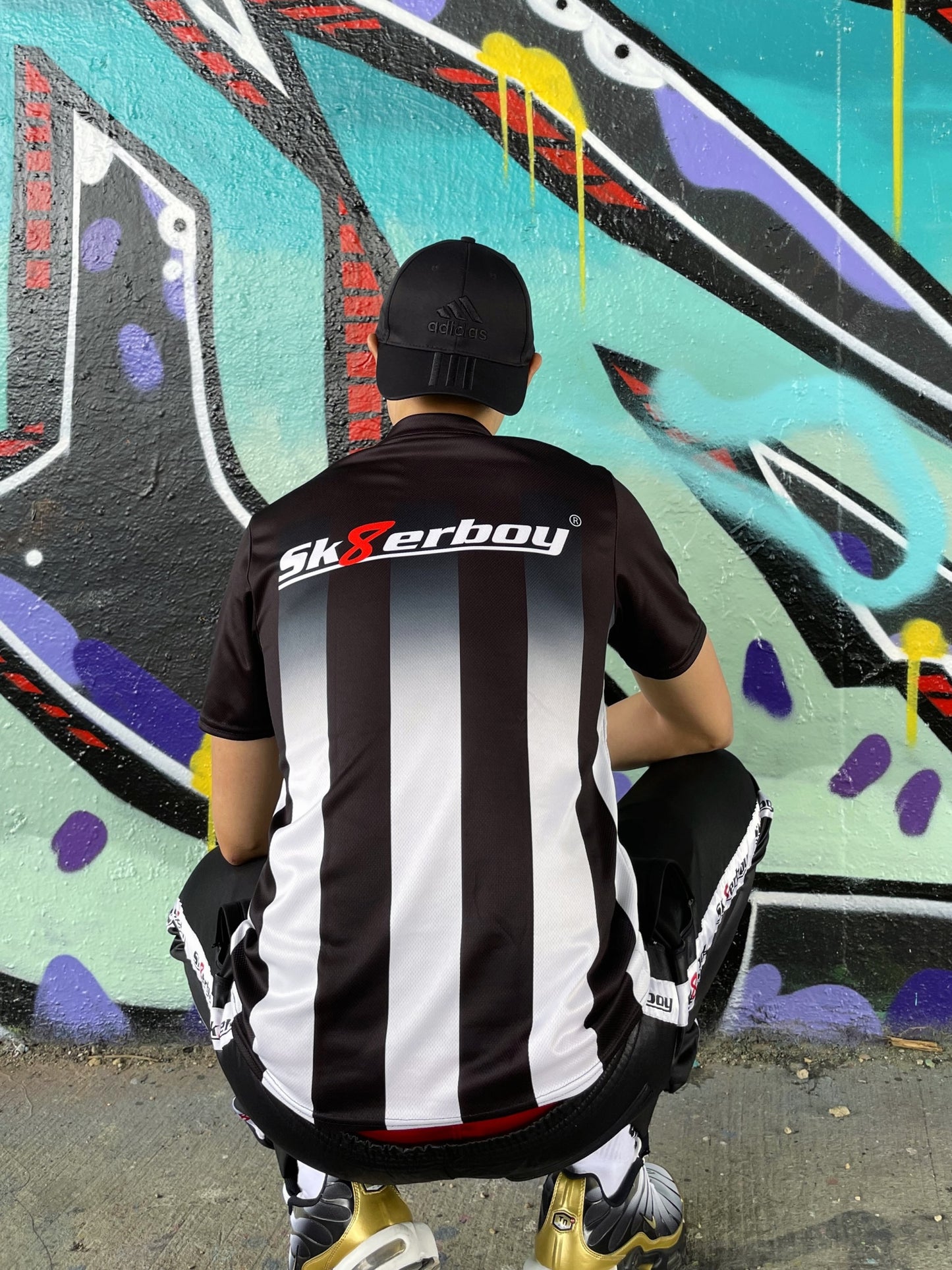 Rückansicht des Sk8erboy® Tikots mit großem Logo, getragen von einem Model in Hocke vor stylischer Graffiti-Kulisse.