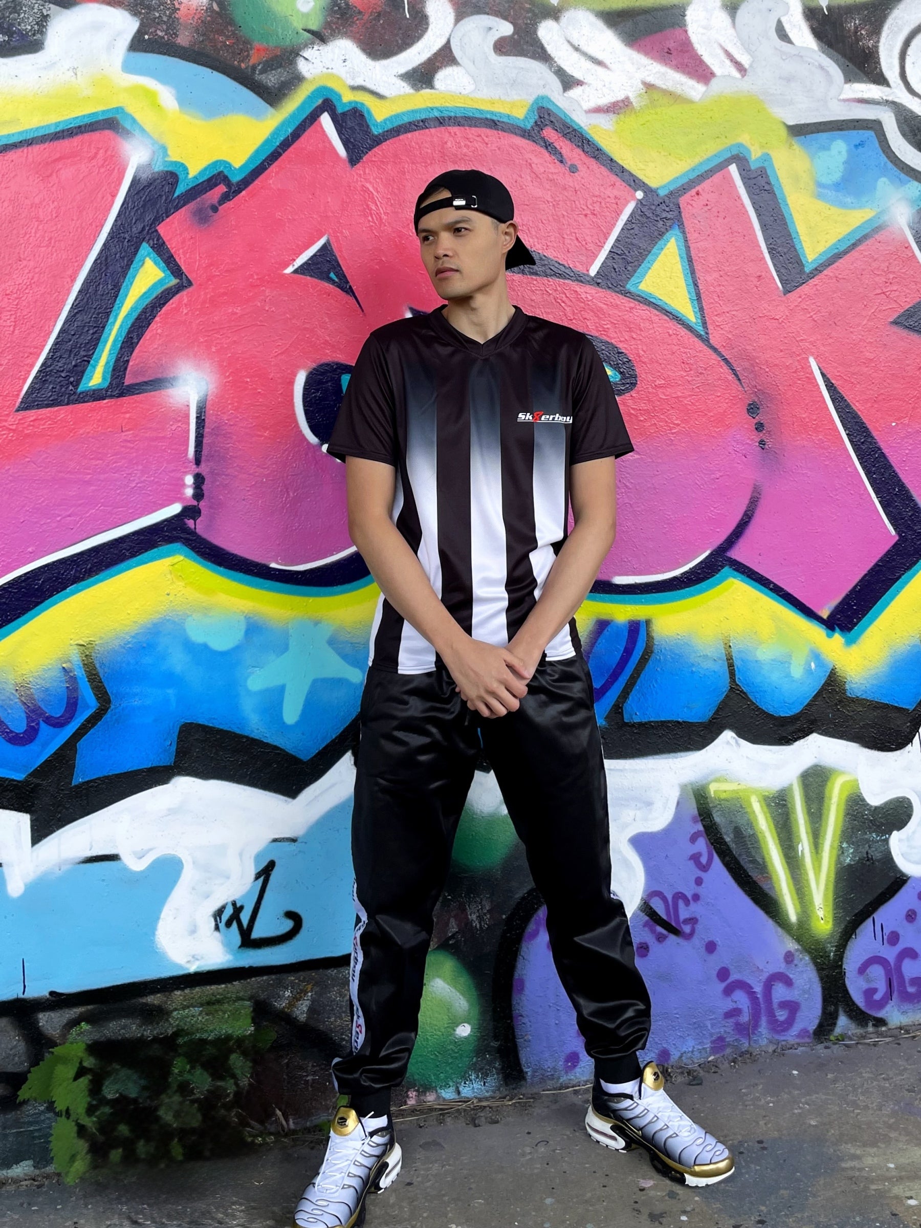 Model trägt das Sk8erboy® Tikot in Schwarz-Weiß mit vertikalen Streifen vor bunter Graffiti-Wand und zeigt auf das Logo auf der Brust.