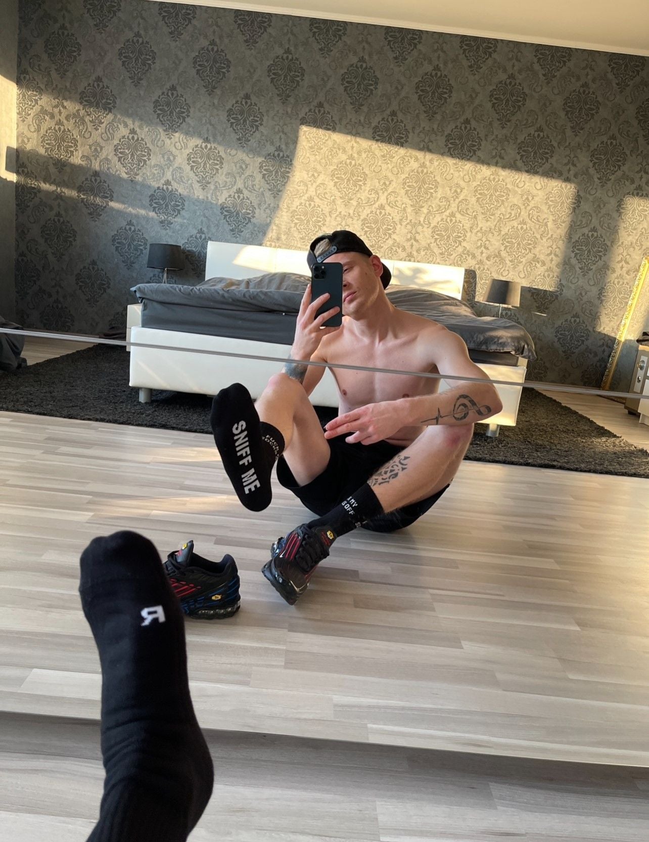 junger typ mit gut gebautem oberkörper im schlafzimmer vor dem bett sitzend zeigt seine schwarzen sniff me socken von sk8erboy in den spiegel und macht ein selfie