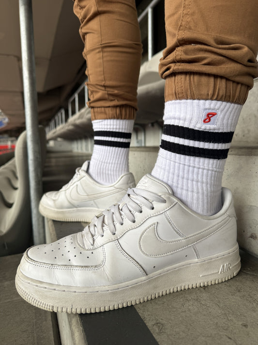 Nahaufnahme von Sk8erboy® Retro Socken mit schwarzen Streifen, getragen mit weißen Nike-Sneakern im Stadion – sportlicher Gay-Look mit Statement-Faktor.