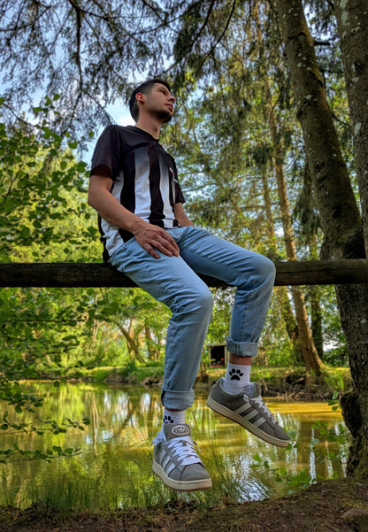 Boy sitzt in der Natur auf einem Holzbalken, trägt hochgekrempelte Jeans, graue Sneaker und weiße Sk8erboy® Socks mit schwarzem Pawprint-Motiv – entspannter Outdoor-Vibe mit Fetisch-Flair.