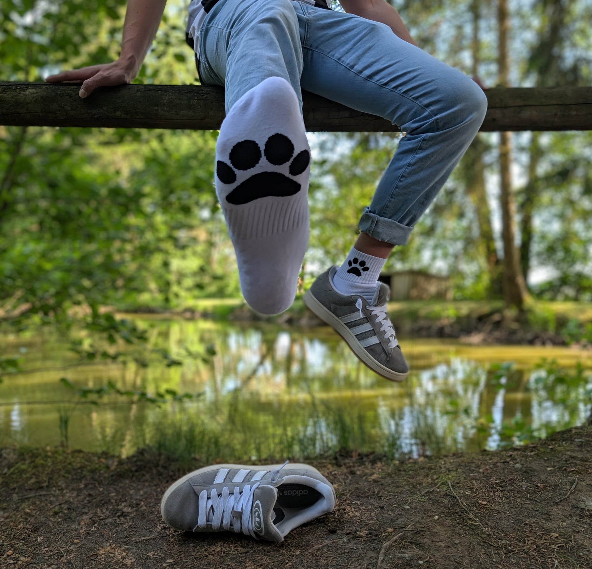 Man sitzt in der Natur auf einem Holzbalken, trägt hochgekrempelte Jeans, graue Sneaker und weiße Sk8erboy® Socks mit schwarzem Pawprint-Motiv – entspannter Outdoor-Vibe mit Fetisch-Flair.
