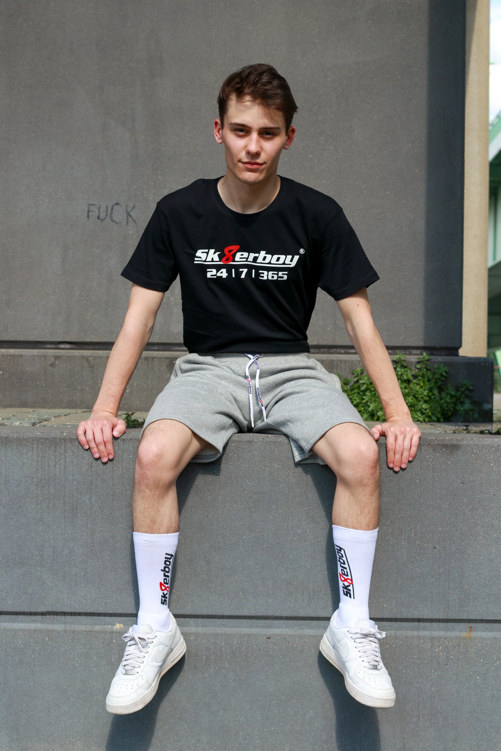 hübscher gay twink trägt ein sk8erboy t-shirt shirt in schwarz baumwolle cotton mit einem sk8erboy aufdruck mit weisser schrift und 8 acht in rot mit 24 7 365 fetter aufdruck mit dem trainingsanzug in kurz und grau von sk8erboy