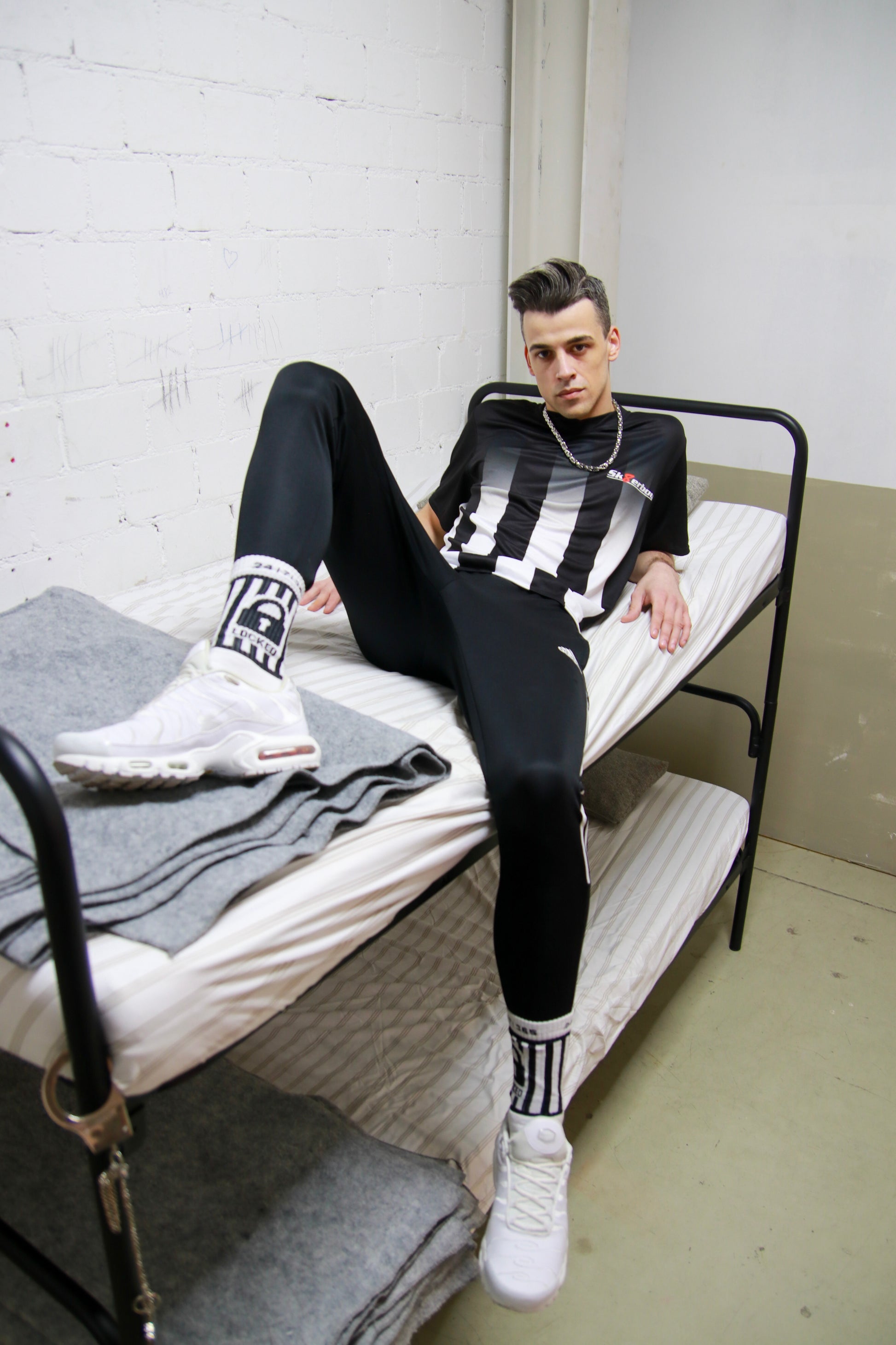 Urbaner Look: Model sitzt mit dem Sk8erboy® Tikot und glänzender Jogginghose in einem Stockbett in einer Gefängniszelle
