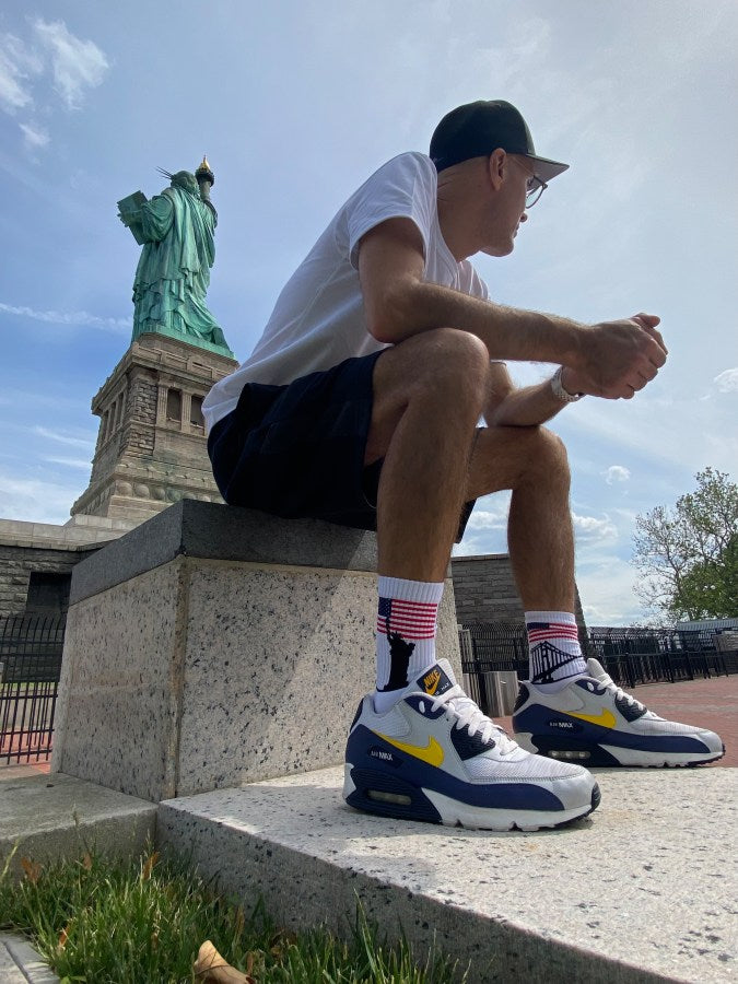 junger sportlicher typ sitzt vor der freiheitsstatue und traegt USA flagge auf weissen socken mit der liberty und golden gate bridge auf dem socken bund  in new york von einem sportlichen jungen getragen mit nike airmax turnschuhen bei sonnenschein in nahaufnahme