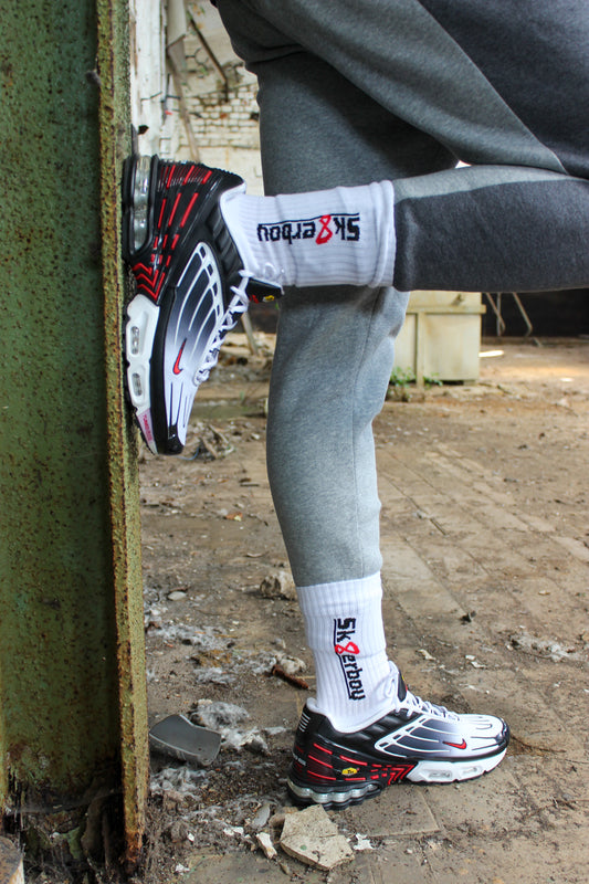 sk8erboy crew socken socks in weiss mit schwarzem schriftzug und roter acht 8 er traegt rot weiss schwarz nike tn tuned 3 drei III er traegt eine graue sweatpant von nike im hintergrund ein lost place farbik