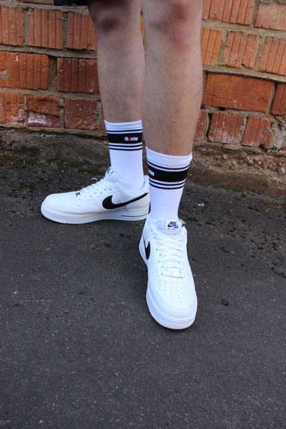 sk8erboy deluxe socken sox in weiss mit schwarzen ringen schwarzen mit schwarzem streifen und logo einstickung rote 8 acht er traegt nike afo air force one low mit weissem leder und schwarzem leder nike swoosh haken  im hintergrund ein lostplace der boy hat einen blanken unterschenkel mit haaren