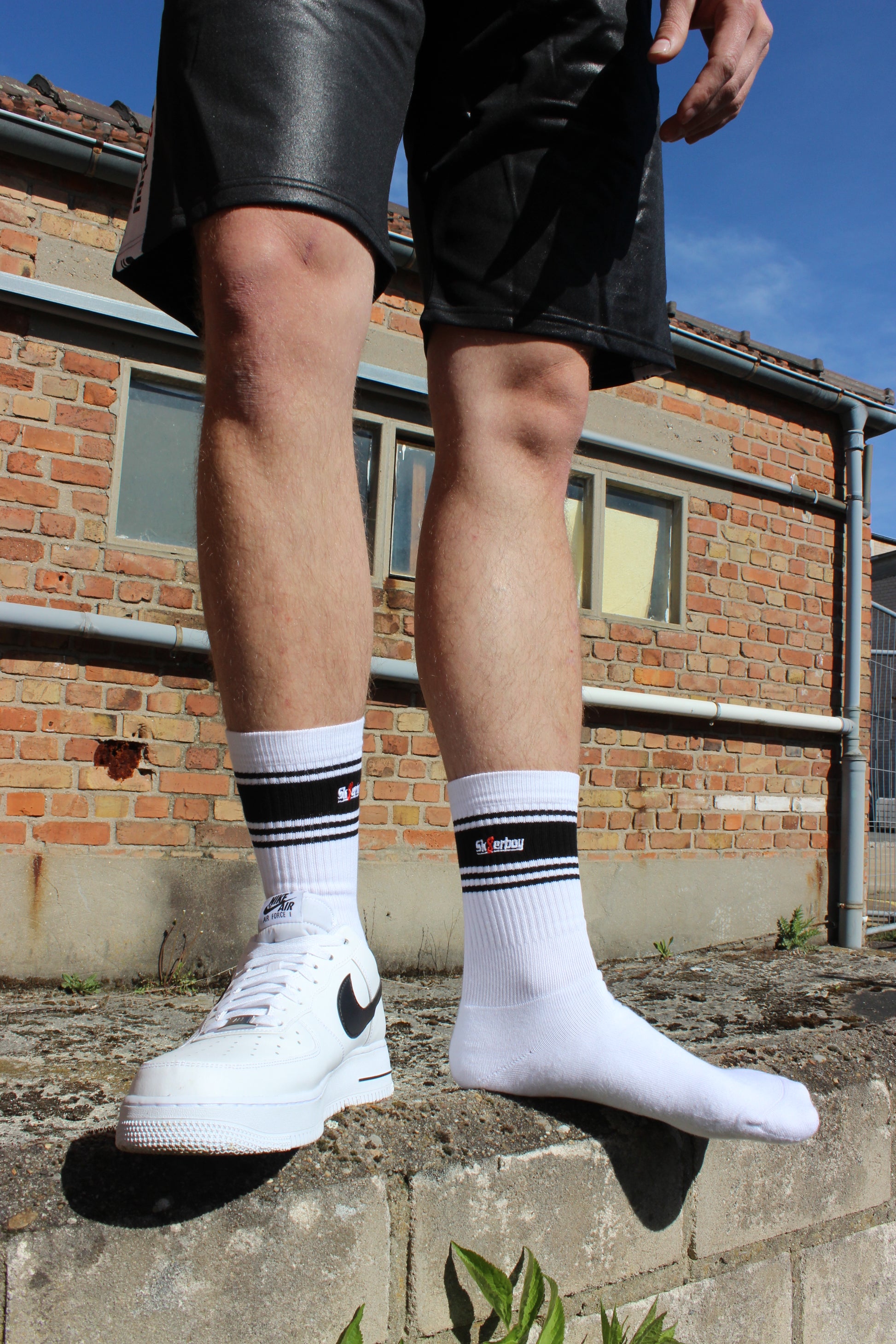 sk8erboy deluxe socken sox in weiss mit schwarzen ringen schwarzen mit schwarzem streifen und logo einstickung rote 8 acht er traegt nike afo air force one low mit weissem leder und schwarzem leder nike swoosh haken  im hintergrund ein lostplace der boy hat einen blanken unterschenkel mit haaren