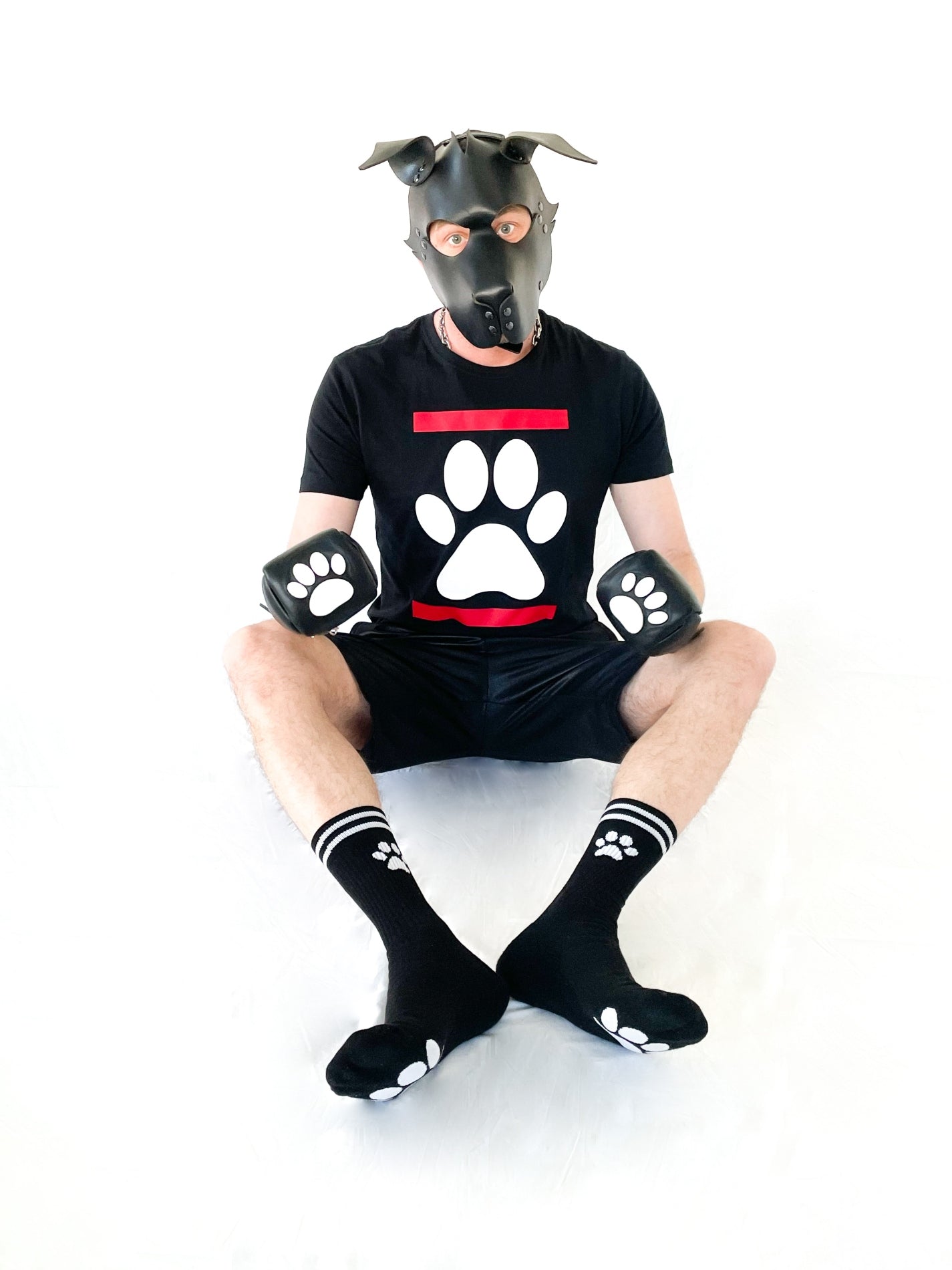 sitzender puppy fetisch mit schwarzen t-shirt und abgebildeter hundepfote auf der vorderseite mit short und schwarzen sk8erboy puppy socken fuer doggys und fetisch spiele