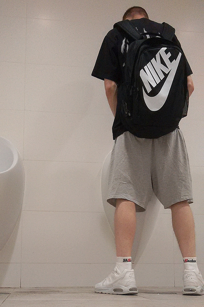 junger gay mit kurzen sportklamotten traegt einen nike rucksack und steht vor einem pissoir mit nike sneaker und sk8erboy quarter socks in weiss