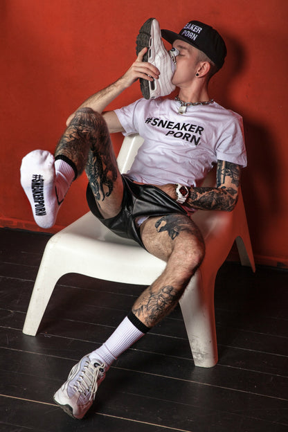 sportlicher junge riecht an seinen turnschuh von nike und trägt eine schwarze boxer berlin short und passende sneakerporn socken von sk8erboy in weiss und schwarz vor einer roten wand sitzend