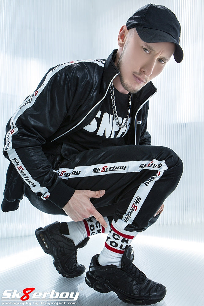 junge in der hocke zeigt auf seine fck you socks von sk8erboy und traegt einen shiny tracksuits trainingsanzug in schwarz mit snffr t-shirt und basecap von nike zu den passenden nike tn sneaker