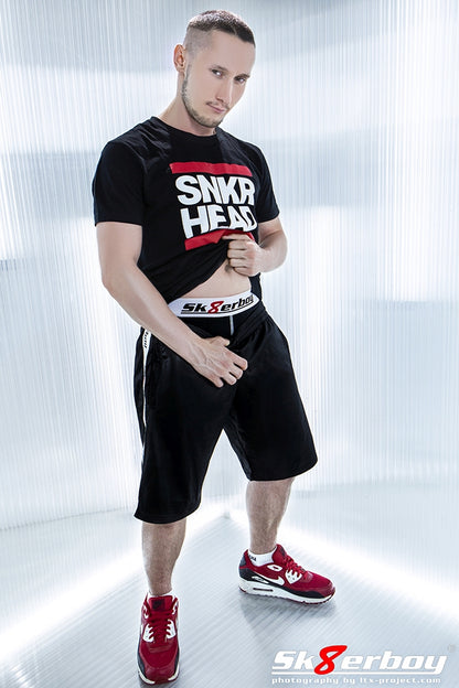 junger typ mit shiny short in schwarz zeigt den bund seiner schwarzen sk8erboy boxershort und traegt passend dazu ein snkr head t-shirt und rote nike air sneaker