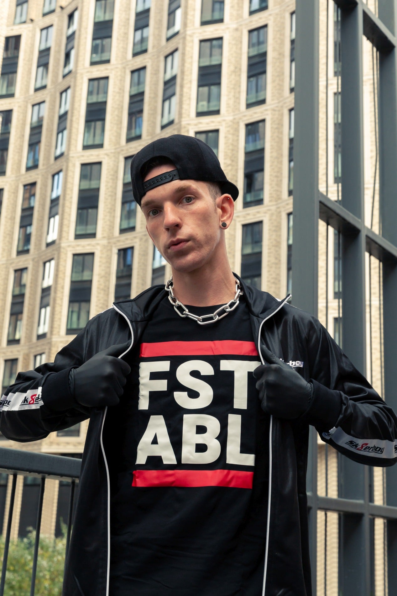 junger gay in der stadt mit schwarzem glanzanzug und t-shirt mit dem aufdruck fst abl traegt schwarze handschuhe und basecap passend zum sk8erboy trainingsanzug