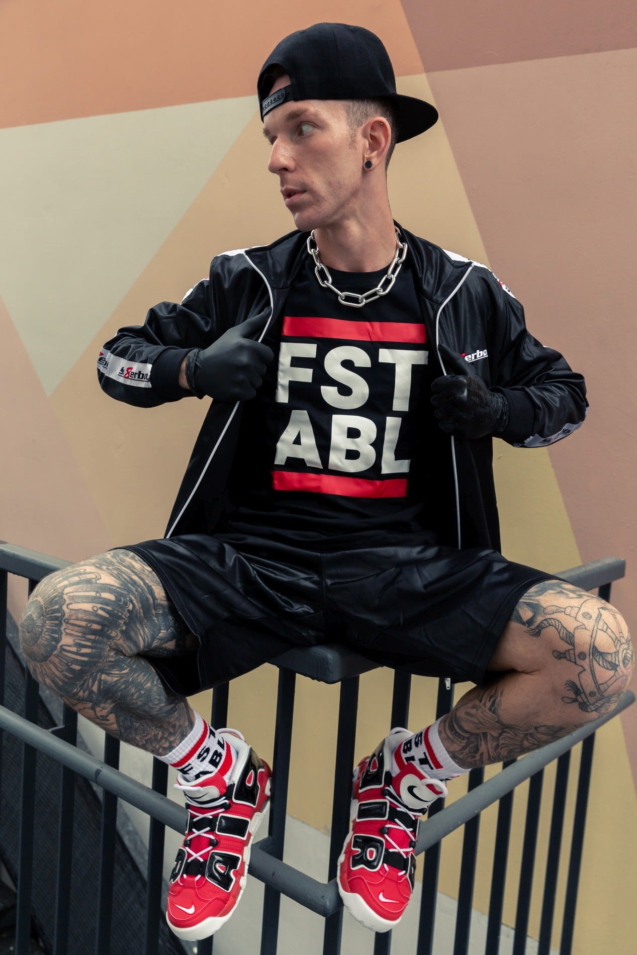 junge sitzt auf einem gelaender mit glanz jacke von sk8erboy und passender short darunter ein schwarzes t-shirt mit fst able auf der vorderseite und passende socken mit nike air sneaks in rot