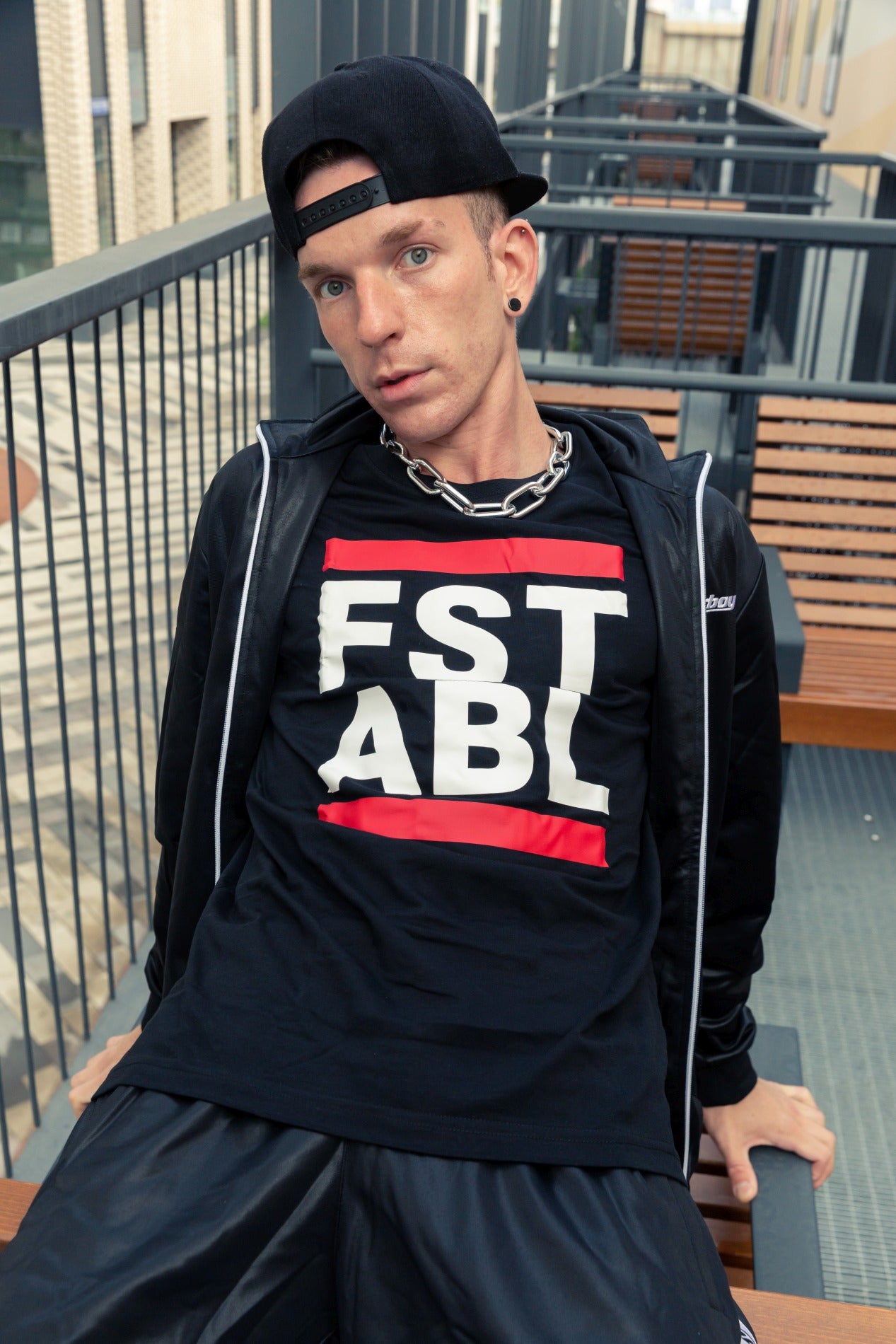 junger boy auf einer terrasse traegt einen shiny trainingsanzug von sk8erboy mit einem schwarzen t-shirt mit dem großen aufdruck fst abl