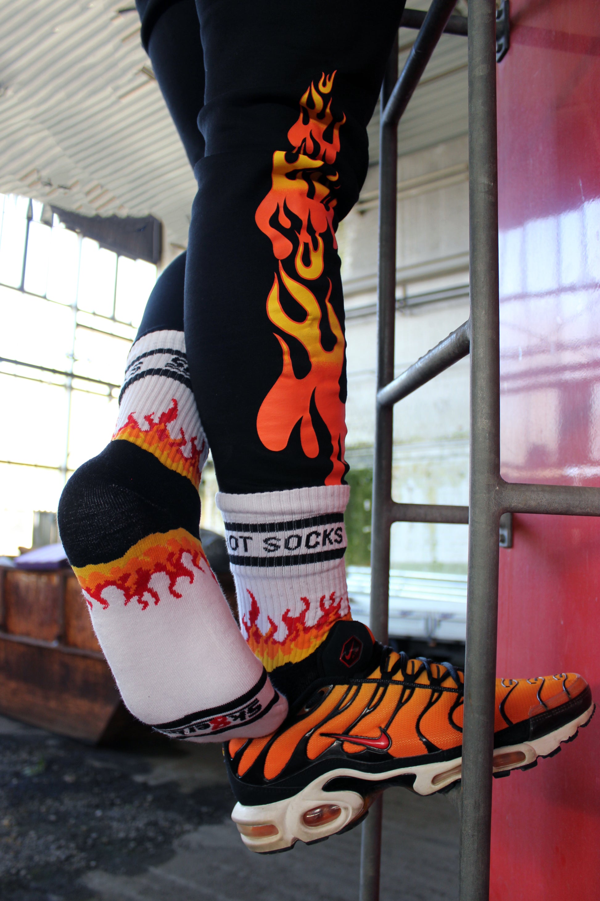sk8erboy hot socks weisse socken mit schwarzem Schriftzug HOT SOCKS in grossen Buchstaben mit einer schwarzen Linie strich oben und unten ansonsten haben die socken ein flammen muster am Schaft und im fussbereich die flammen sind rot orange und gelb mit einer schwarzen Absetzung der boy Kerl traegt tiger nike tn tuned mit einem roten swosh haken er steht auf einer Leiter die an einem Feuerwehr auto steht