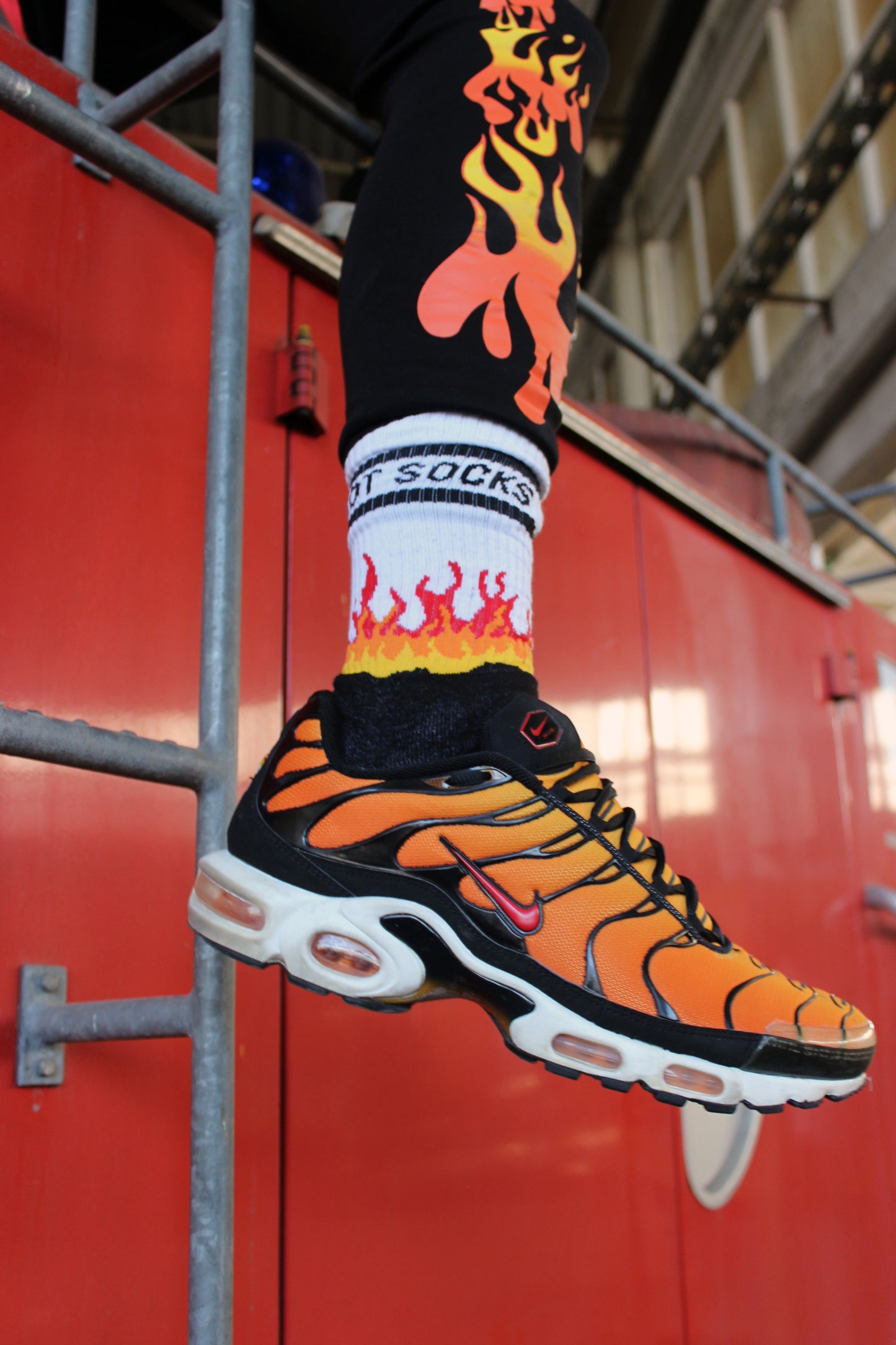 sk8erboy hot socks weisse socken mit schwarzem Schriftzug HOT SOCKS in grossen Buchstaben mit einer schwarzen Linie strich oben und unten ansonsten haben die socken ein flammen muster am Schaft und im fussbereich die flammen sind rot orange und gelb mit einer schwarzen Absetzung der boy Kerl traegt tiger nike tn tuned mit einem roten swosh haken er steht auf einer Leiter die an einem Feuerwehr auto steht