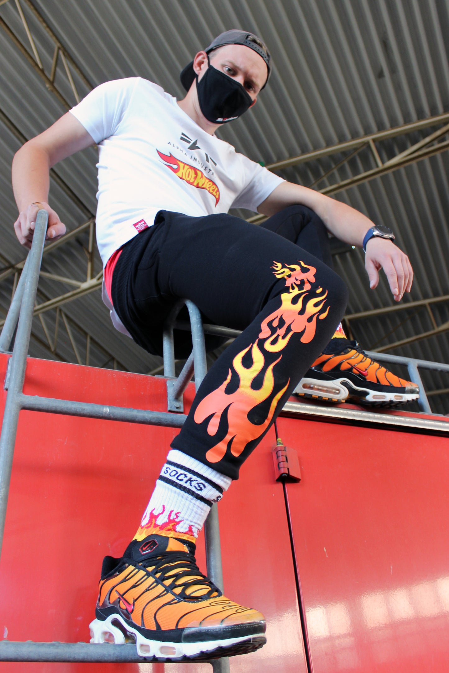 sk8erboy hot socks weisse socken mit schwarzem Schriftzug HOT SOCKS in grossen Buchstaben mit einer schwarzen Linie strich oben und unten ansonsten haben die socken ein flammen muster am Schaft und im fussbereich die flammen sind rot orange und gelb mit einer schwarzen Absetzung der boy Kerl traegt tiger nike tn tuned mit einem roten swosh haken er steht auf einer Leiter die an einem Feuerwehr auto steht man sieht noch eine die schwarze alpha industries hose mit der hot wheels Fahne und einer Mund