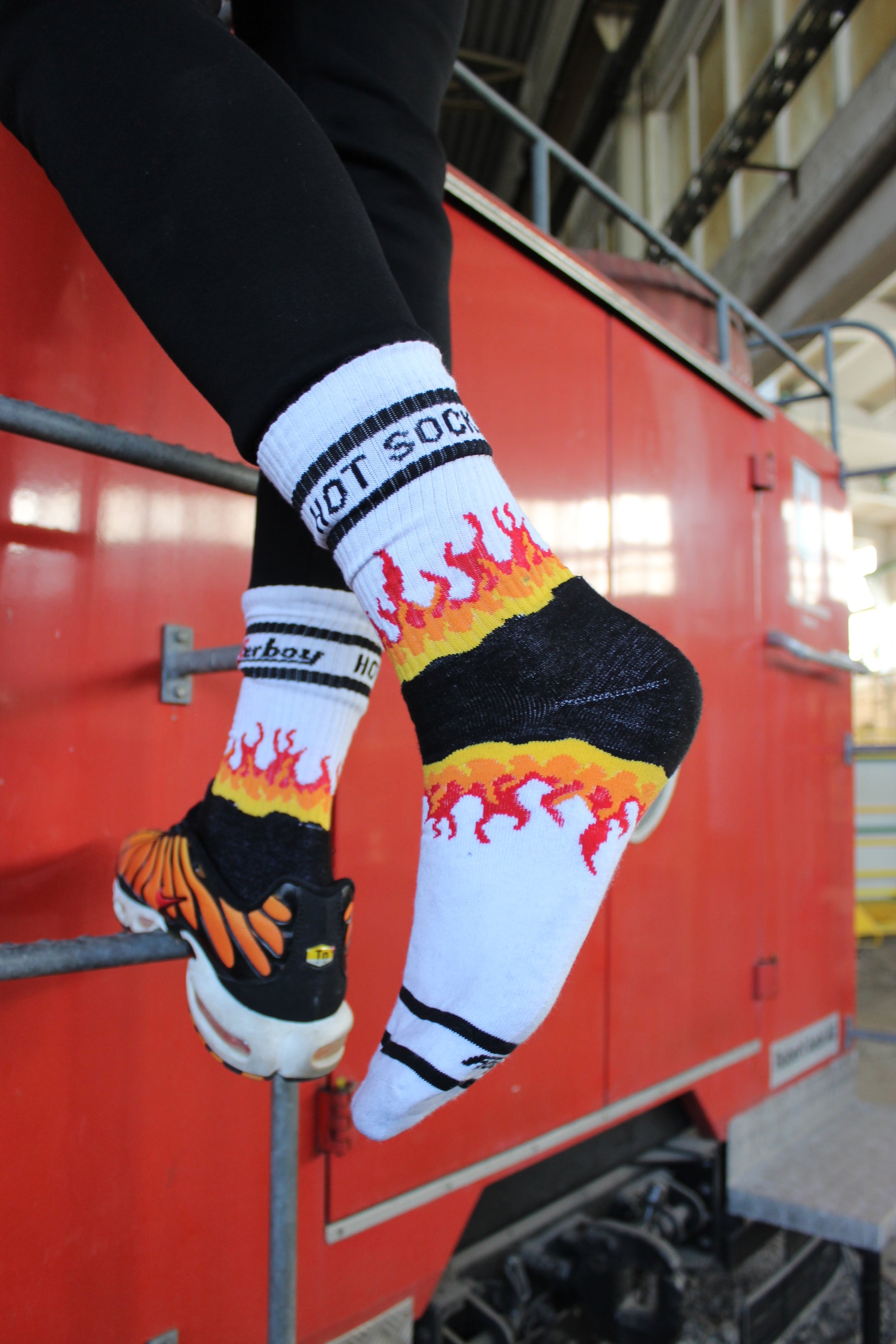 sk8erboy hot socks weisse socken mit schwarzem Schriftzug HOT SOCKS in grossen Buchstaben mit einer schwarzen Linie strich oben und unten ansonsten haben die socken ein flammen muster am Schaft und im fussbereich die flammen sind rot orange und gelb mit einer schwarzen Absetzung der boy Kerl traegt tiger nike tn tuned mit einem roten swosh haken er steht auf einer Leiter die an einem Feuerwehr auto steht mauer haengt