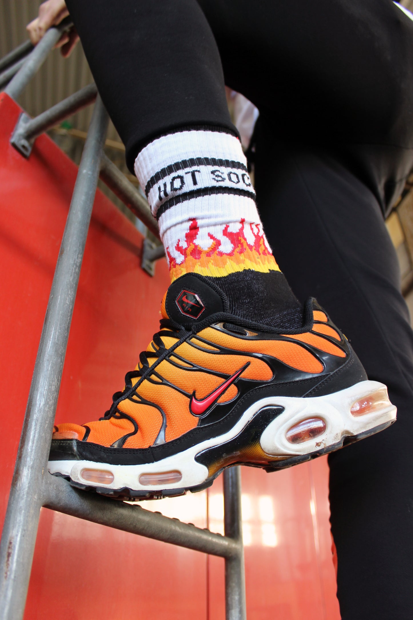 sk8erboy hot socks weisse socken mit schwarzem Schriftzug HOT SOCKS in grossen Buchstaben mit einer schwarzen Linie strich oben und unten ansonsten haben die socken ein flammen muster am Schaft und im fussbereich die flammen sind rot orange und gelb mit einer schwarzen Absetzung der boy Kerl traegt tiger nike tn tuned mit einem roten swosh haken er steht auf einer Leiter die an einem Feuerwehr auto steht