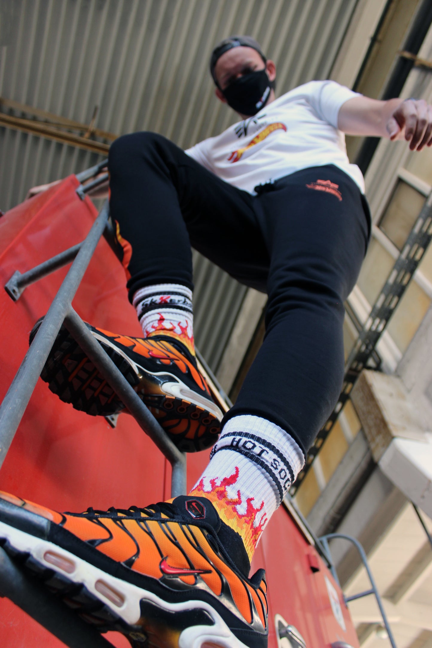 sk8erboy hot socks weisse socken mit schwarzem Schriftzug HOT SOCKS in grossen Buchstaben mit einer schwarzen Linie strich oben und unten ansonsten haben die socken ein flammen muster am Schaft und im fussbereich die flammen sind rot orange und gelb mit einer schwarzen Absetzung der boy Kerl traegt tiger nike tn tuned mit einem roten swosh haken er steht auf einer Leiter die an einem Feuerwehr auto steht man sieht noch eine die schwarze alpha industries hose mit der hot wheels Fahne und einer Mund