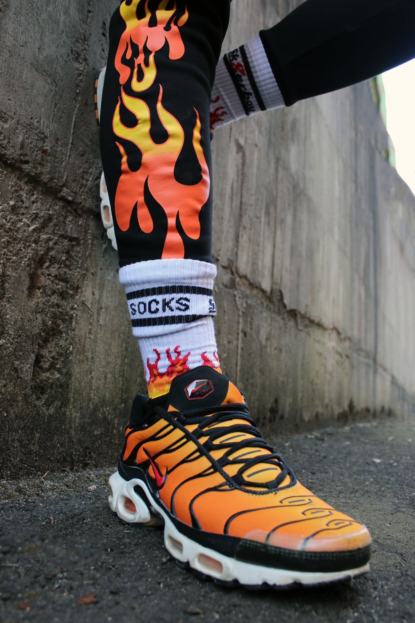 sk8erboy hot socks weisse socken mit schwarzem Schriftzug HOT SOCKS in grossen Buchstaben mit einer schwarzen Linie strich oben und unten ansonsten haben die socken ein flammen muster am Schaft und im fussbereich die flammen sind rot orange und gelb mit einer schwarzen Absetzung der boy Kerl traegt tiger nike tn tuned mit einem roten swosh haken er steht auf einer Leiter die an einem Feuerwehr auto steht mauer haengt