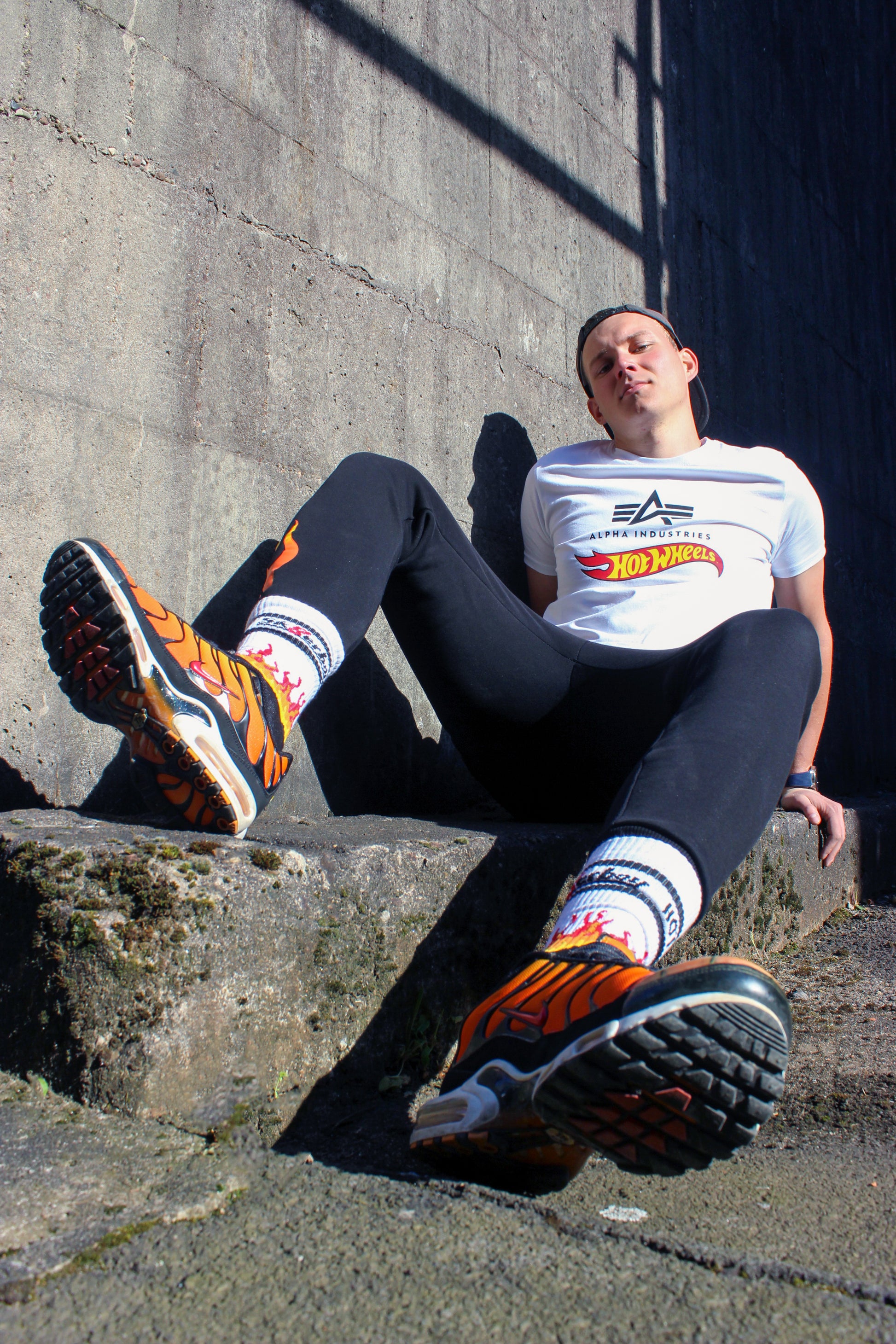 der boy liegt am Boden mit einem geilen blick er hat eine cap basecap auf dem kopf er traegt ein weisses alpha industries t-shirt shirt mit einem schwarzen Aufdruck und dem rot gelben hot wheels logo weiter hat er eine jogger pant an in schwarz auf mir dem logo seine feet Fuesse sind flauschig verpackt in sk8erboy hot socks mit flammen und smelly Absetzungen die nike tn tuned air max plus im tiger style stehen dampfen neben ihm willig zu sniffen