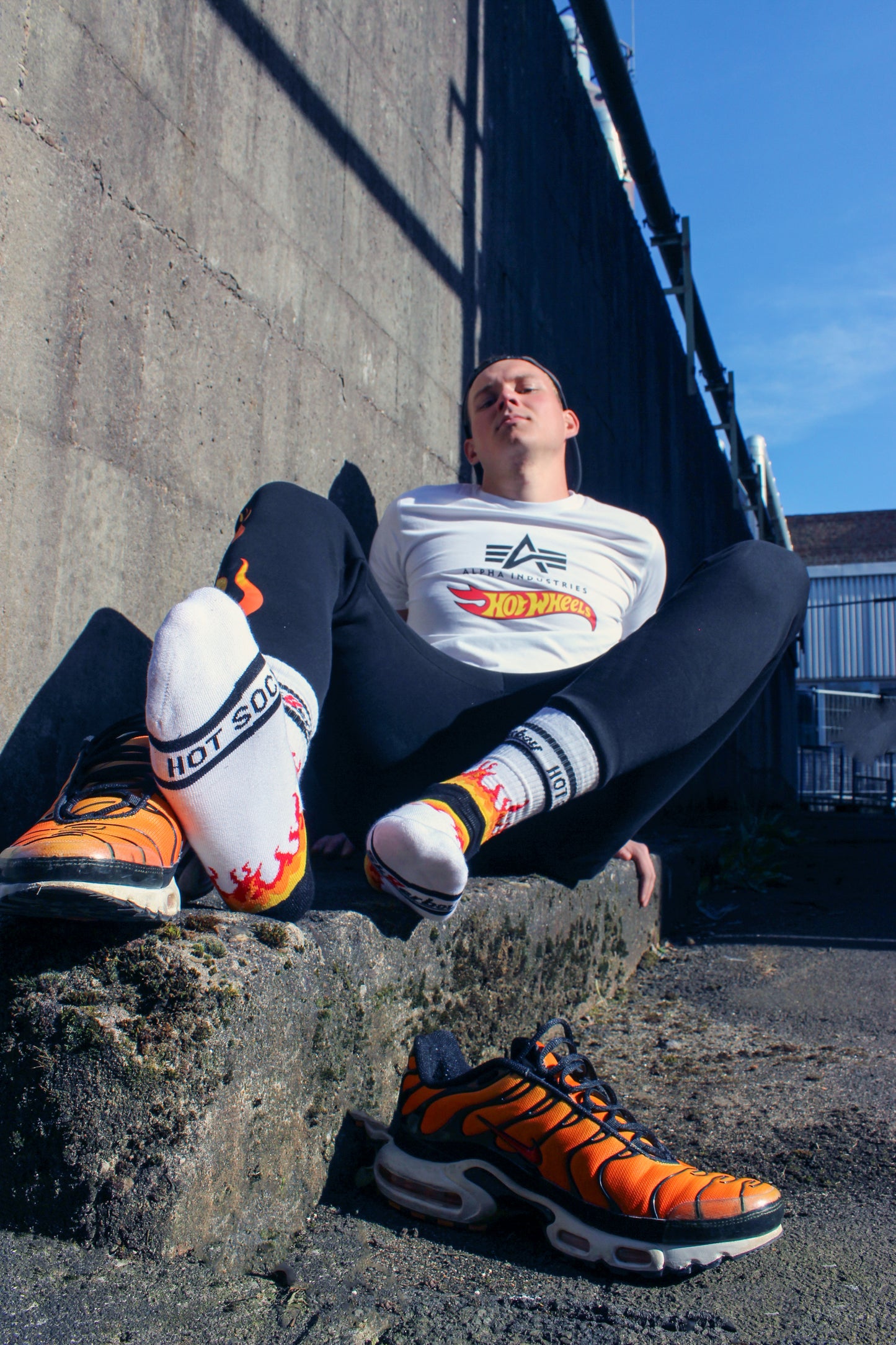 der boy liegt am Boden mit einem geilen blick er hat eine cap basecap auf dem kopf er traegt ein weisses alpha industries t-shirt shirt mit einem schwarzen Aufdruck und dem rot gelben hot wheels logo weiter hat er eine jogger pant an in schwarz auf mir dem logo seine feet Fuesse sind flauschig verpackt in sk8erboy hot socks mit flammen und smelly Absetzungen die nike tn tuned air max plus im tiger style stehen dampfen neben ihm willig zu sniffen