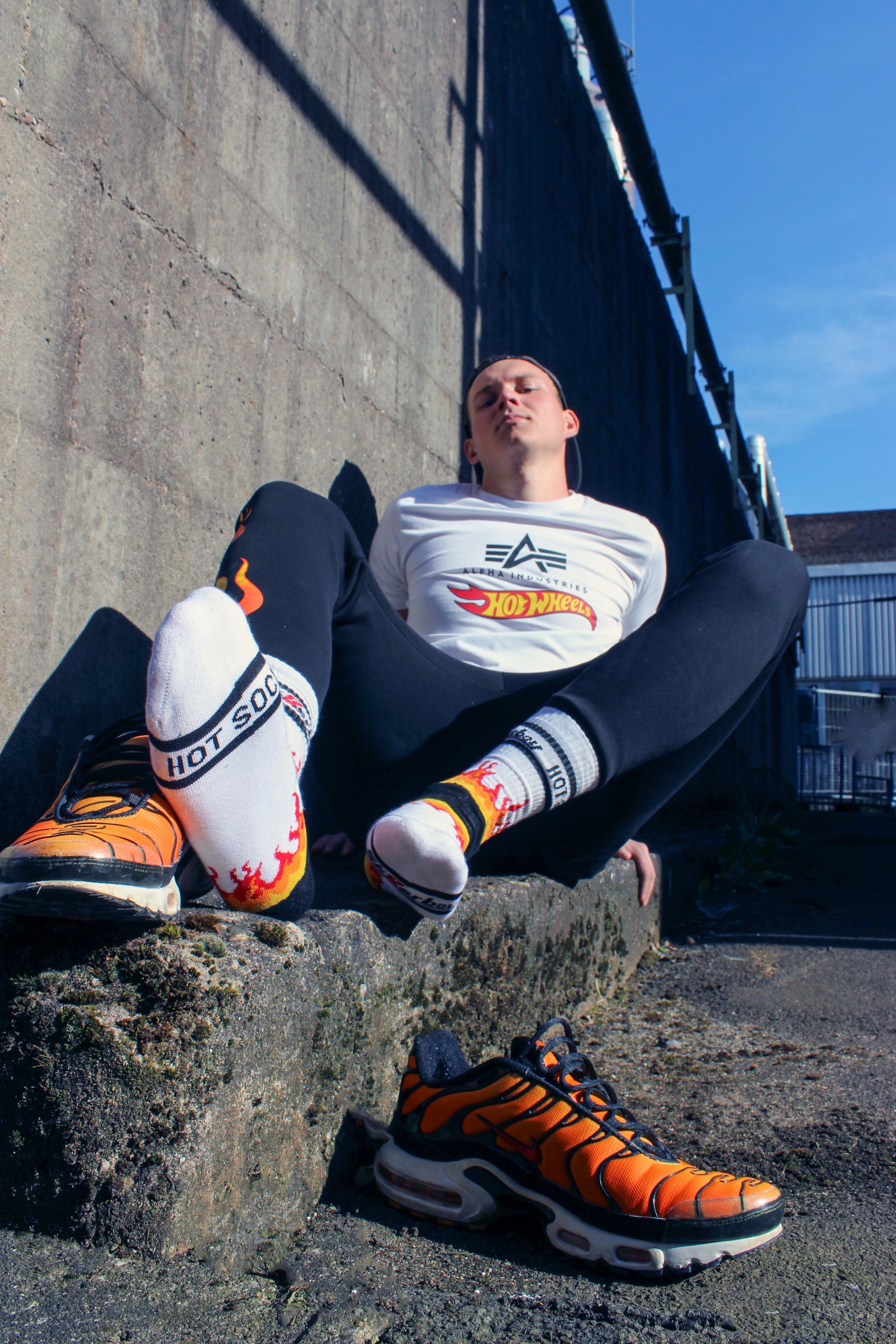 der boy liegt am Boden mit einem geilen blick er hat eine cap basecap auf dem kopf er traegt ein weisses alpha industries t-shirt shirt mit einem schwarzen Aufdruck und dem rot gelben hot wheels logo weiter hat er eine jogger pant an in schwarz auf mir dem logo seine feet Fuesse sind flauschig verpackt in sk8erboy hot socks mit flammen und smelly Absetzungen die nike tn tuned air max plus im tiger style stehen dampfen neben ihm willig zu sniffen