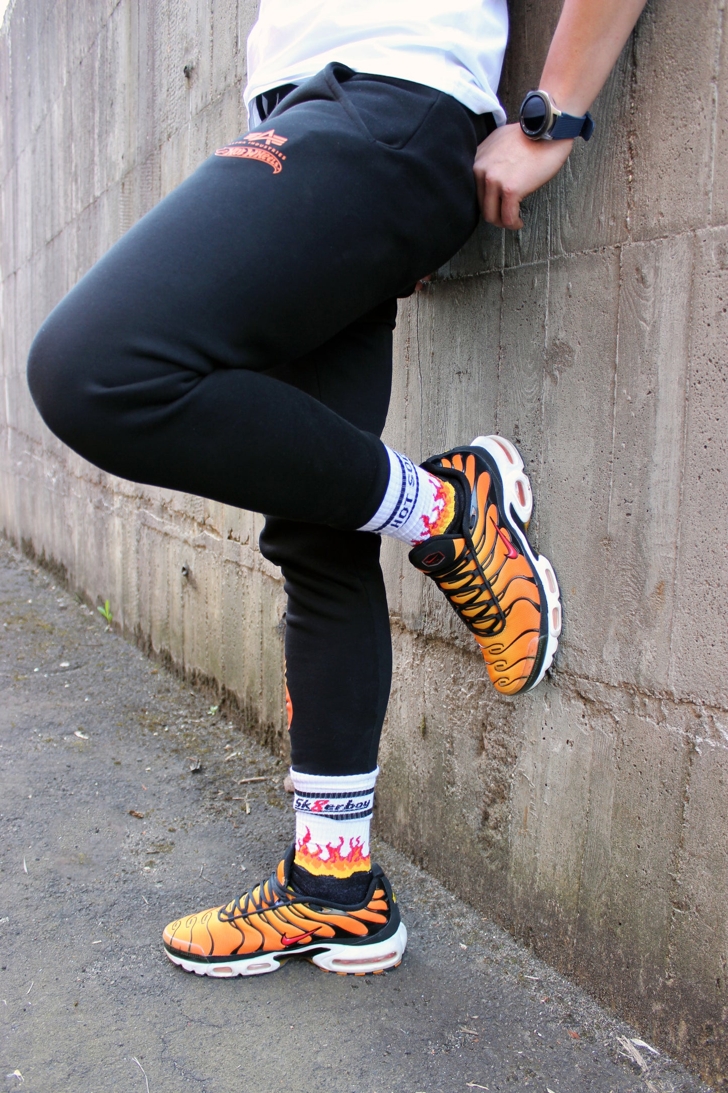 sk8erboy hot socks weisse socken mit schwarzem Schriftzug HOT SOCKS in grossen Buchstaben mit einer schwarzen Linie strich oben und unten ansonsten haben die socken ein flammen muster am Schaft und im fussbereich die flammen sind rot orange und gelb mit einer schwarzen Absetzung der boy Kerl traegt tiger nike tn tuned mit einem roten swosh haken er steht auf einer Leiter die an einem Feuerwehr auto steht mauer haengt