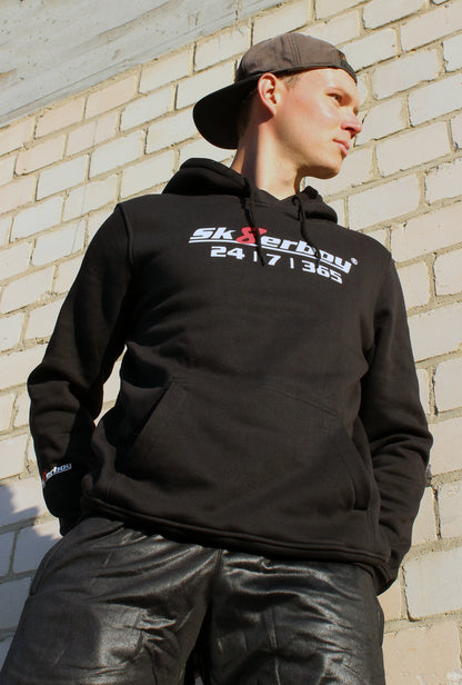 boy traegt einen sk8erboy kapuzen pullover mit einem aufdruck sk8erboy 365 7 24 mit einem eindeutigen slogan ebenso sk8erboy acht 8 in rot mit weissem schriftzug auf der brust und dem aermel der boy traegt eine cap im hintergrund ein lost place und eine shiny sk8erboy short pant kurze hose